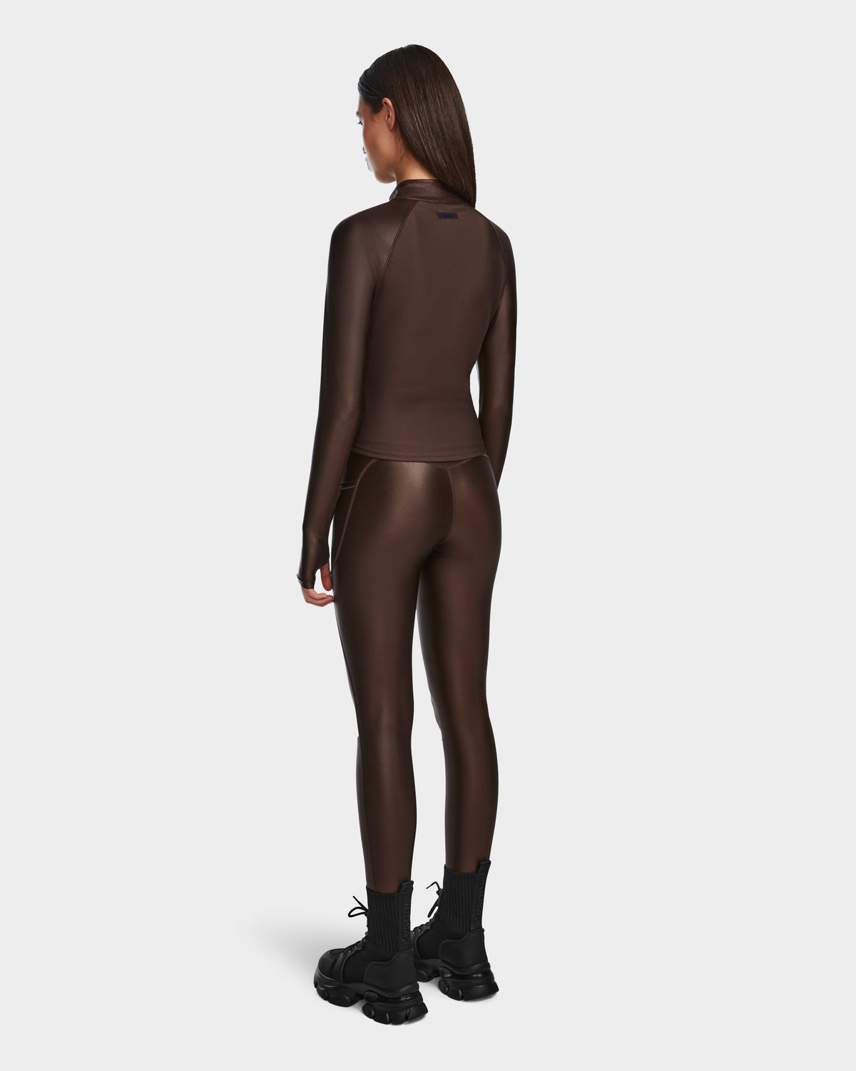 A woman faces away, wearing the Rudsak EVA - 8524514 Espresso fitted long-sleeve zip-up top and matching shiny stretch leggings with black ankle boots against a plain white background.