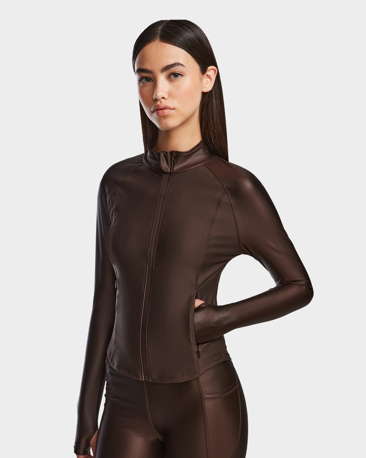 A woman with straight dark hair poses confidently against a plain background, wearing the shiny stretch EVA - 8524514 Espresso zip-up top and matching pants by Rudsak, with one hand on her hip.
