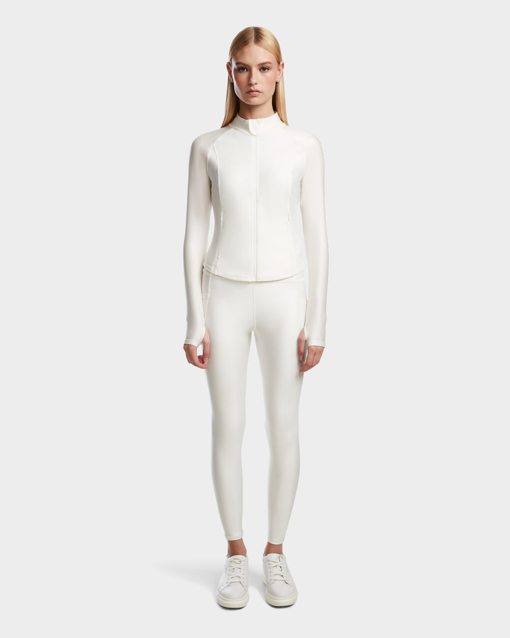A woman faces forward against a light gray background, wearing the Rudsak EVA - 8524514 Iceland fitted long-sleeve zip-up top with matching white shiny stretch leggings and white sneakers.