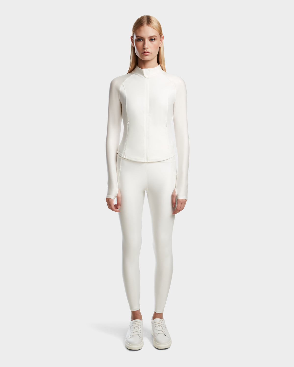 A woman faces forward against a light gray background, wearing the Rudsak EVA - 8524514 Iceland fitted long-sleeve zip-up top with matching white shiny stretch leggings and white sneakers.