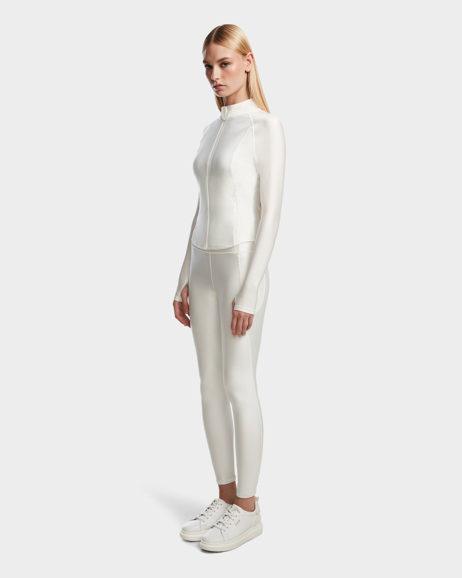A woman with straight blonde hair models the Rudsak EVA - 8524514 Iceland fitted long-sleeve women’s zip-up top, featuring a waterproof zipper, paired with matching white pants and sneakers against a plain light background.
