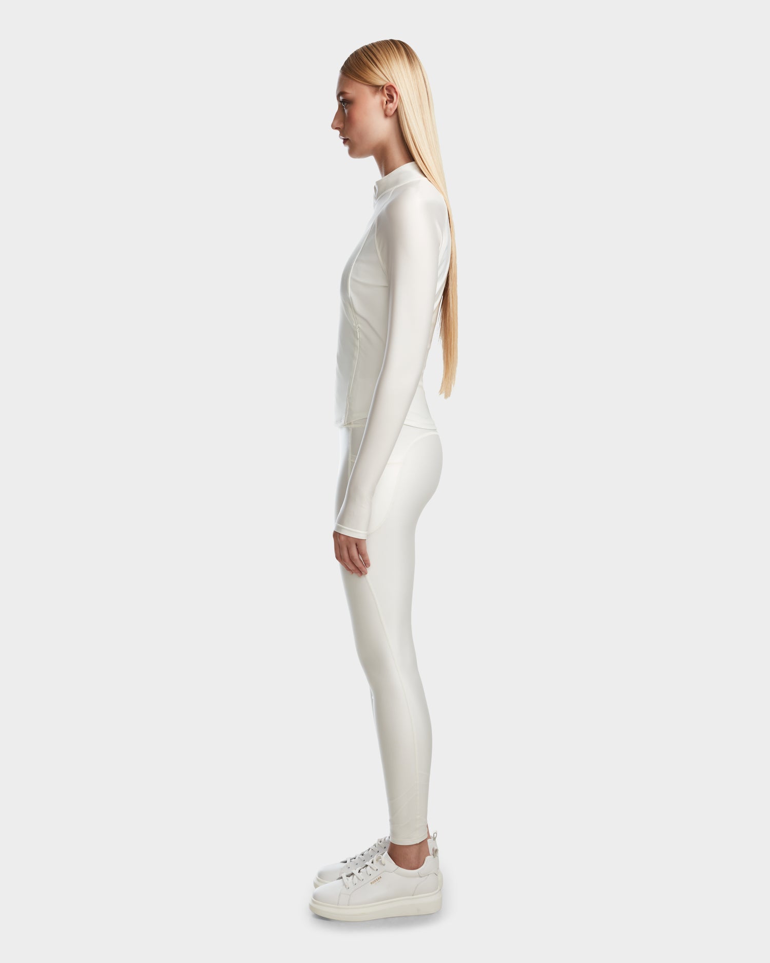 A woman with long blonde hair stands in profile wearing Rudsak’s EVA - 8524514 Iceland fitted white zip-up top and matching leggings, paired with platform sneakers against a plain light background.