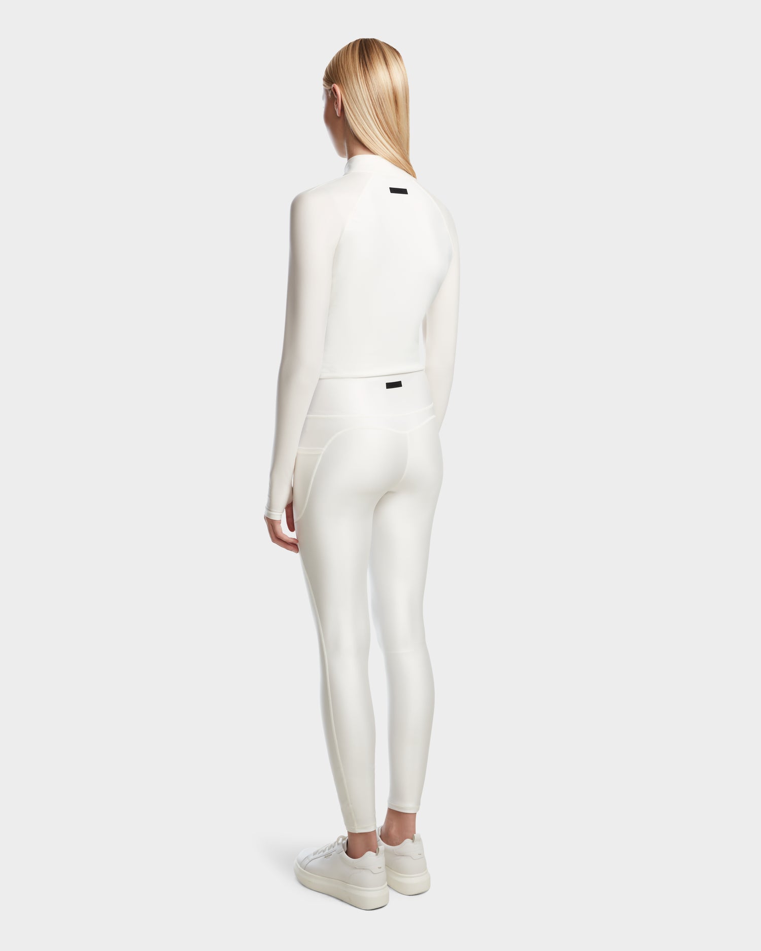 A woman with long blonde hair wears the Rudsak EVA - 8524514 Iceland fitted zip-up top and matching high-waisted leggings in shiny stretch fabric, paired with white sneakers. She stands facing away against a plain light gray background.