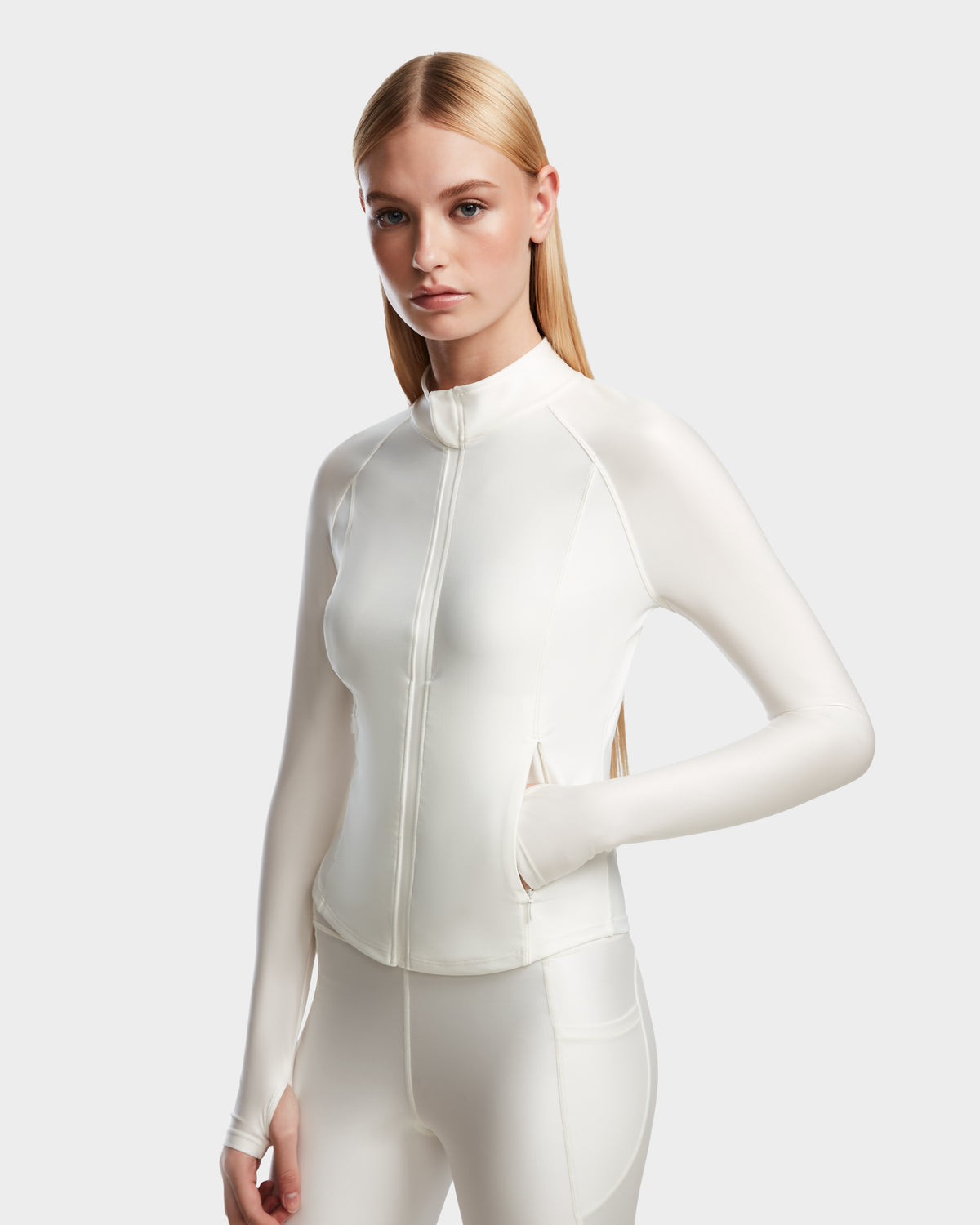 A woman with long blonde hair wears the Rudsak EVA - 8524514 Iceland womens fitted zip-up top and matching shiny stretch white leggings, standing with one hand in her pocket against a plain light background.