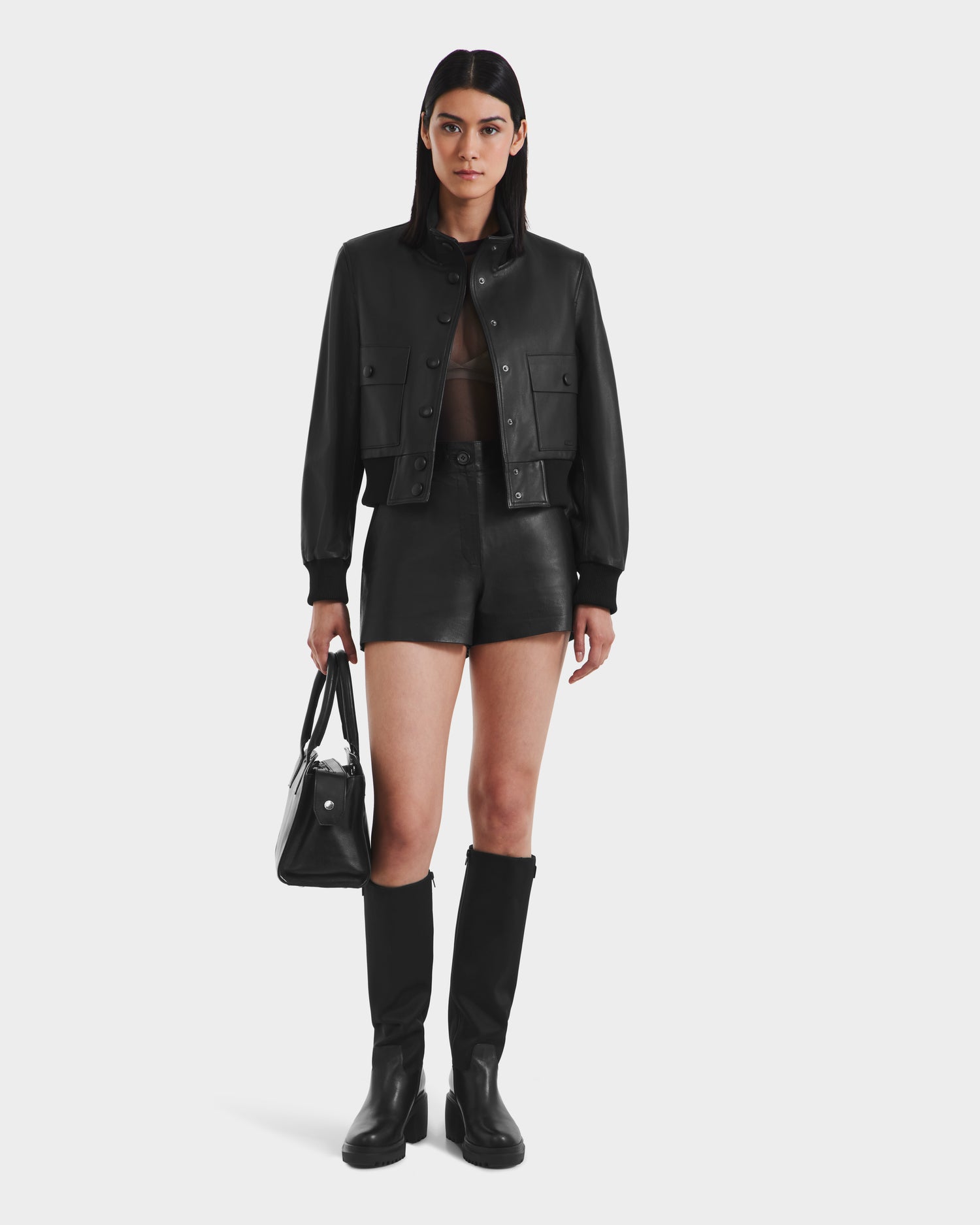 A woman faces forward, wearing the EVERLEIGH - 8125547 Black leather bomber jacket by Rudsak with black shorts, knee-high boots, and a black handbag. She has straight hair and a neutral expression against a plain white background.