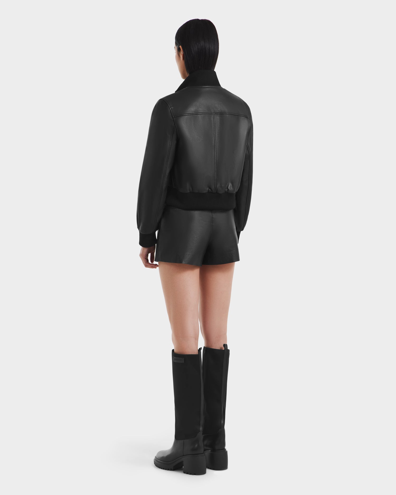 A person stands facing away in the Rudsak EVERLEIGH - 8125547 Black womens leather bomber jacket with matching black leather shorts and tall black boots against a plain white background.