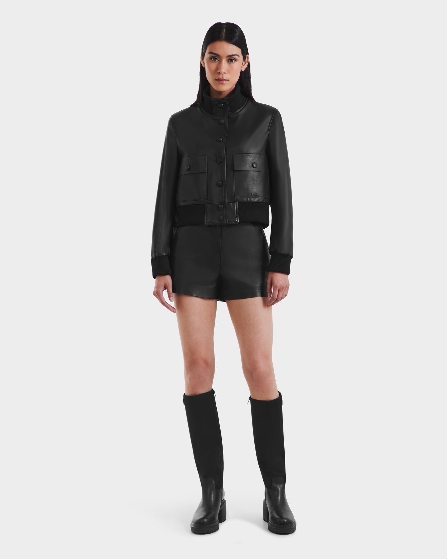 A woman stands facing forward in the Rudsak EVERLEIGH - 8125547 Black leather bomber jacket with black leather shorts and knee-high black boots against a plain white background.