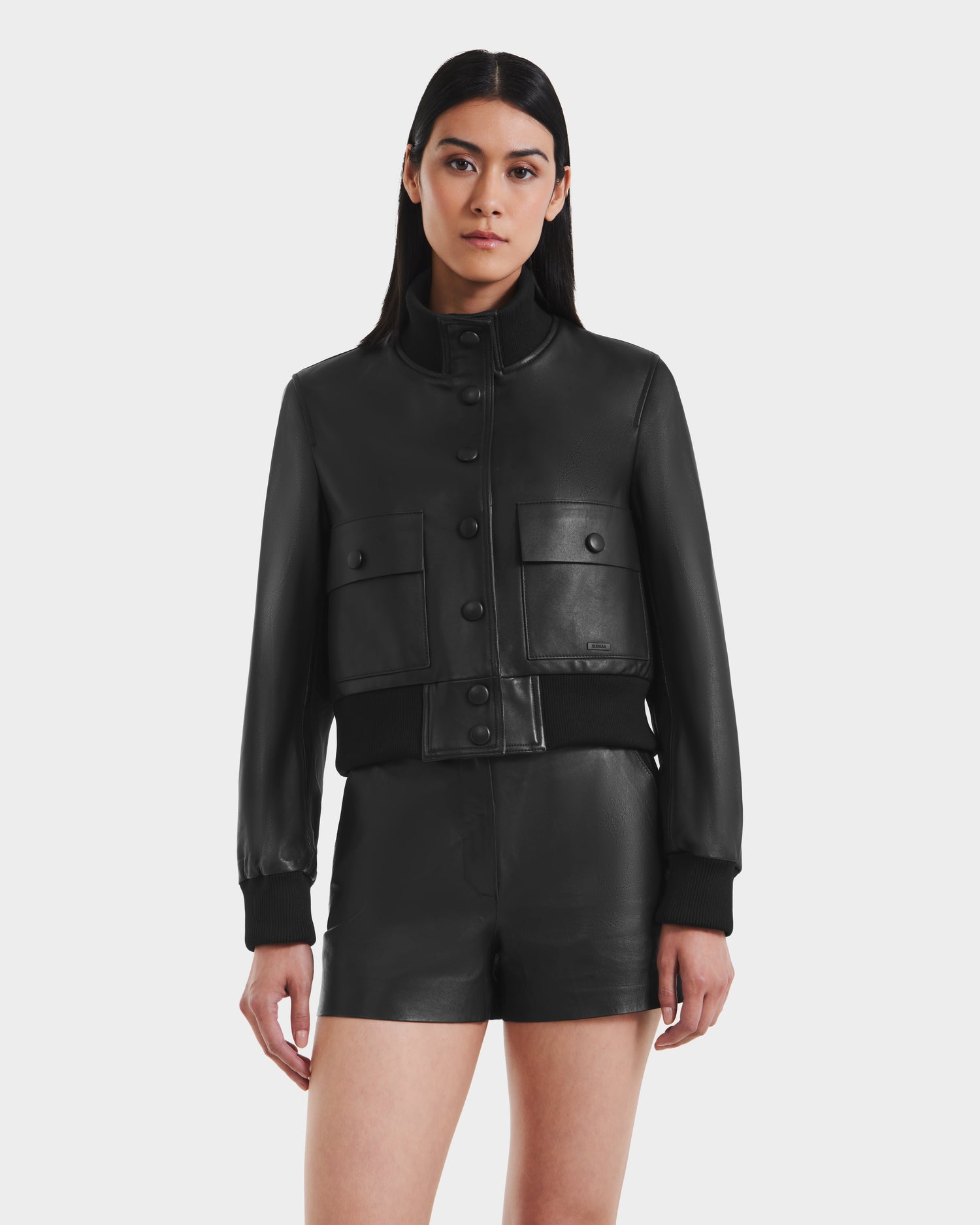 A woman with long dark hair wears the Rudsak EVERLEIGH - 8125547 Black leather bomber jacket with front pockets and matching black leather shorts, standing against a plain white background.