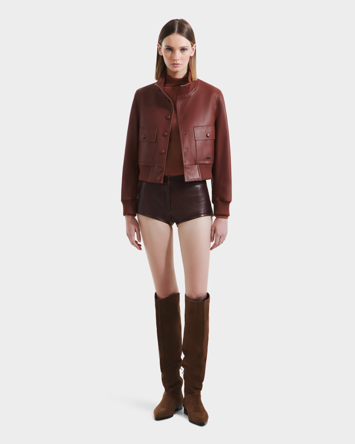 A woman stands facing forward wearing the Rudsak EVERLEIGH - 8125547 Rust leather bomber jacket, paired with a matching brown top, dark brown leather shorts, and tall brown suede boots against a plain white background.