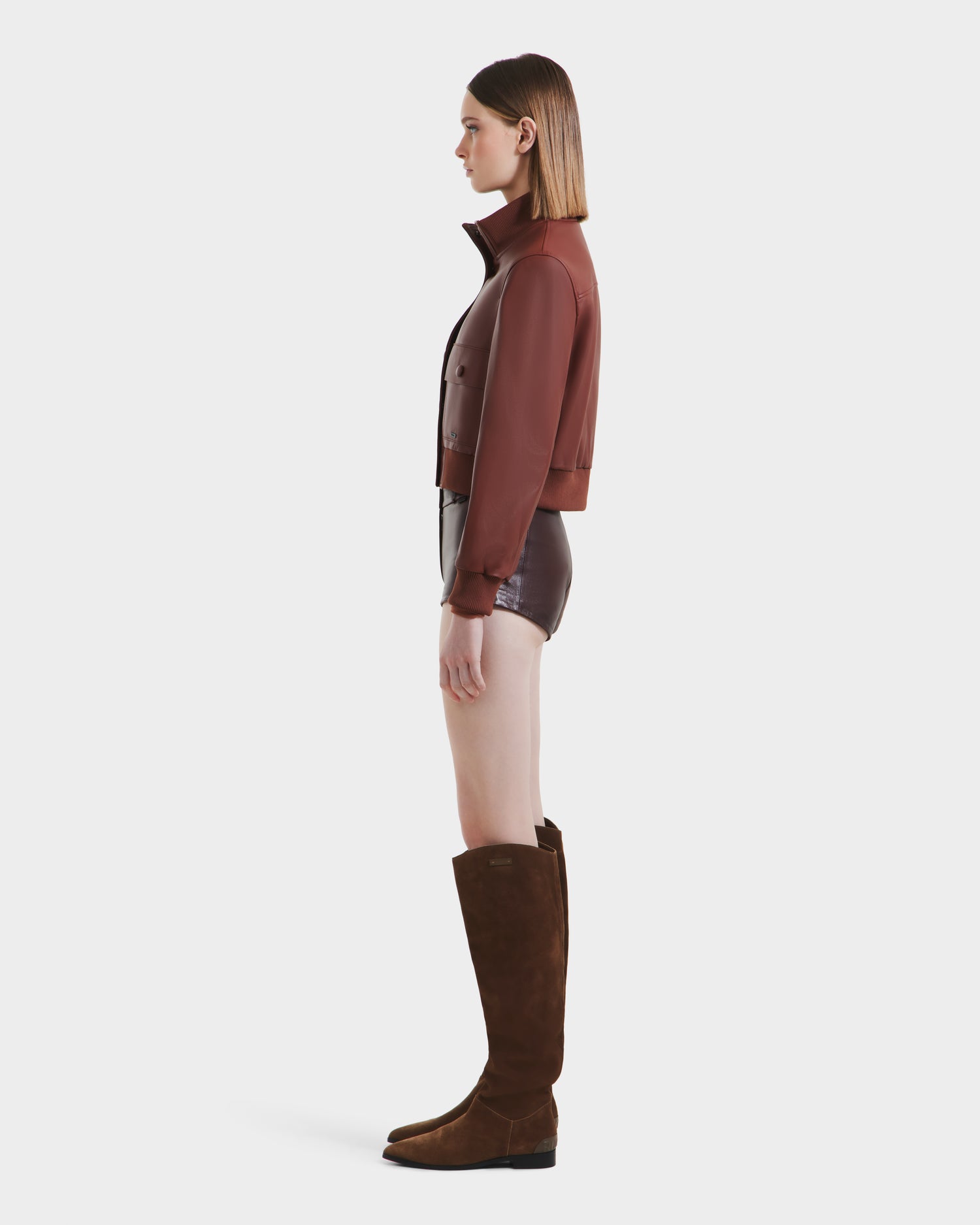 A woman stands in profile against a white background, wearing the Rudsak EVERLEIGH - 8125547 Rust leather bomber jacket with glossy brown shorts and knee-high brown suede boots. Her straight, shoulder-length hair is tucked behind her ears.