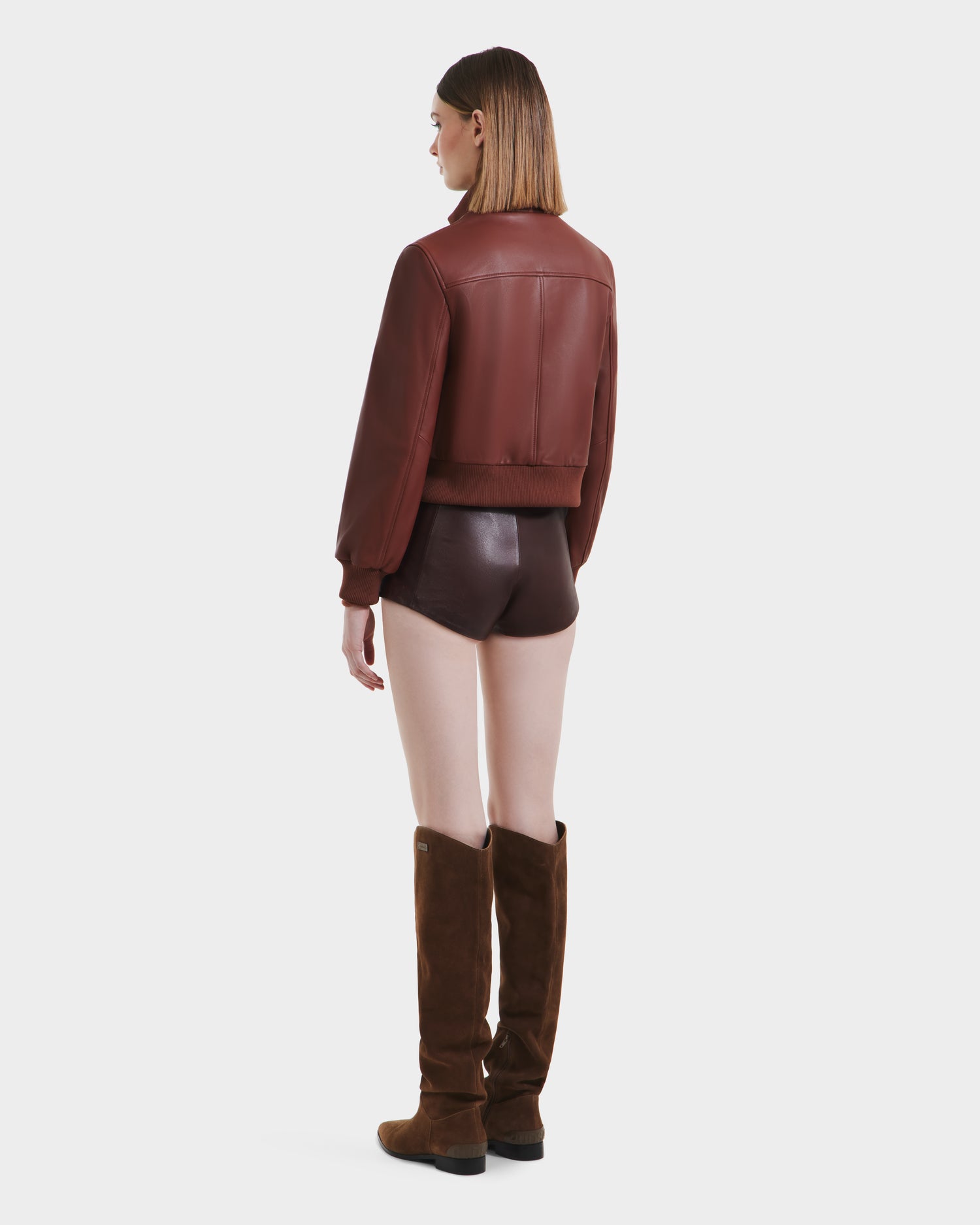A woman faces away, wearing the Rudsak EVERLEIGH - 8125547 Rust leather bomber jacket with matching brown leather shorts and tall brown suede boots against a plain white background.