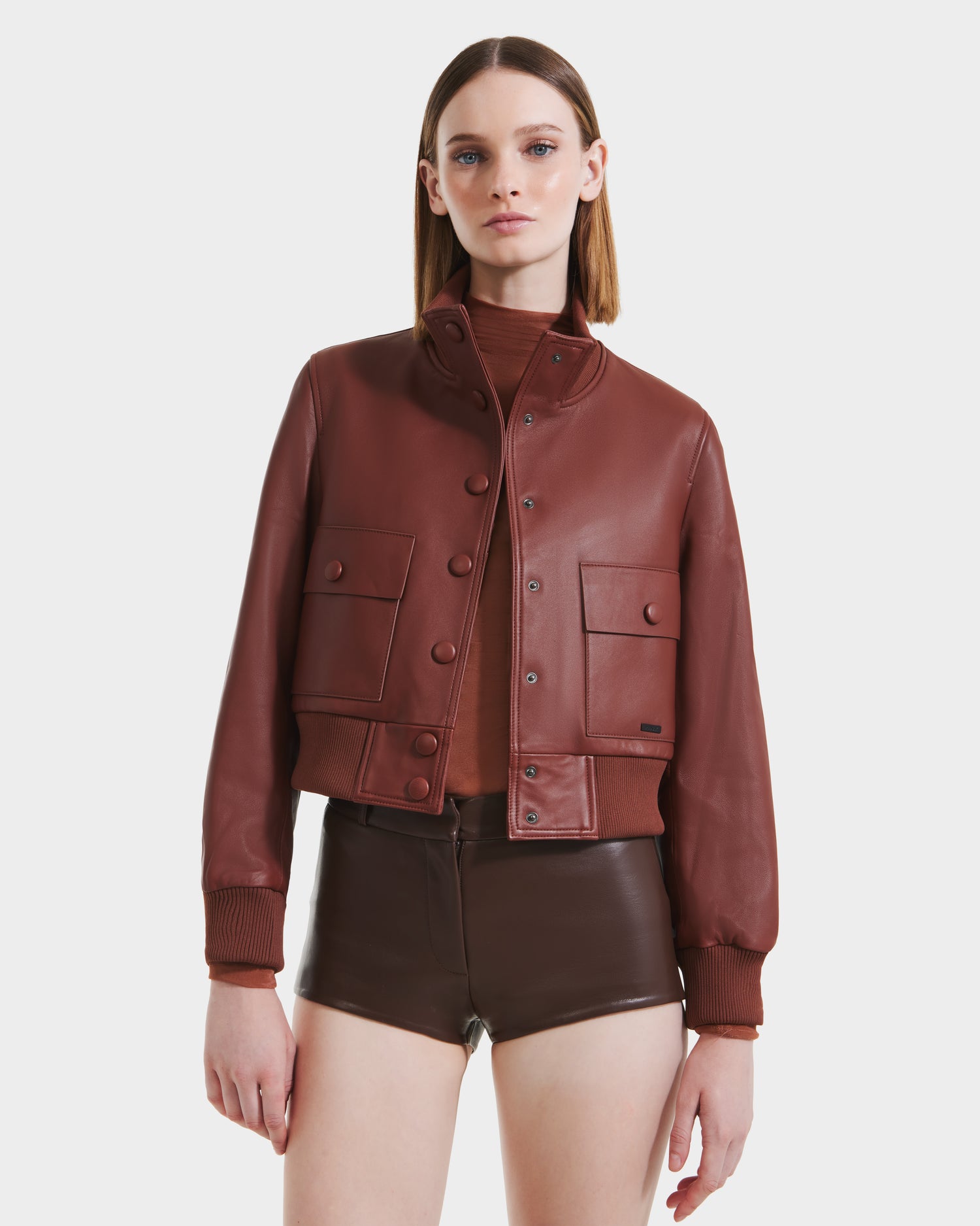 A woman faces forward, wearing the Rudsak EVERLEIGH - 8125547 Rust leather bomber jacket over a brown top and matching brown leather shorts against a plain white background.