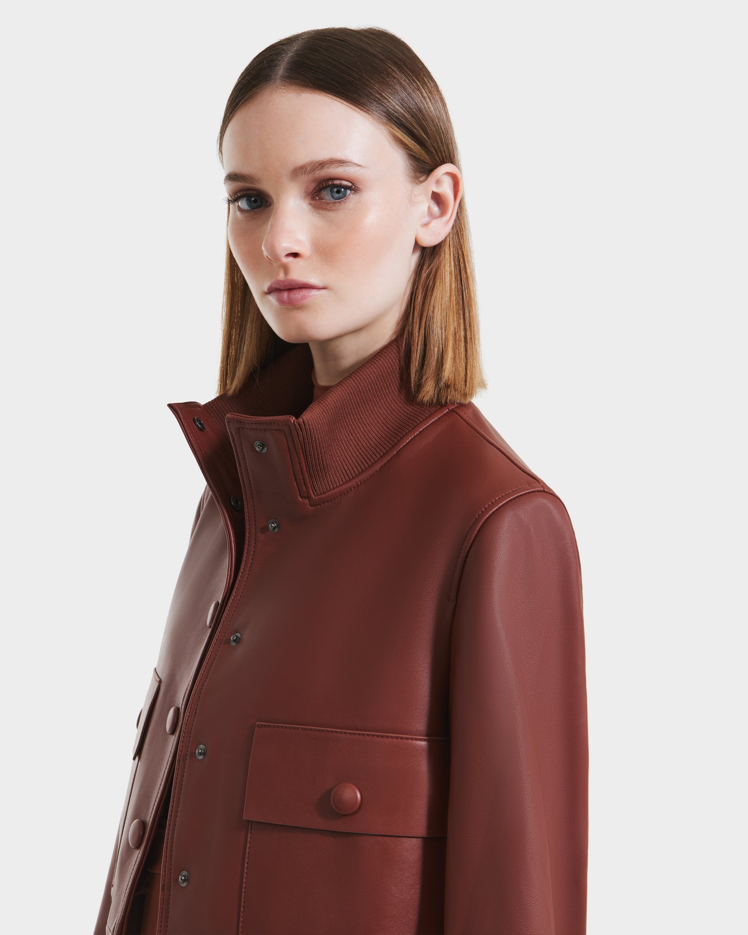 A woman with straight, shoulder-length brown hair wears the Rudsak EVERLEIGH - 8125547 Rust women’s leather bomber jacket, featuring a high collar and large front buttons, standing against a plain white background.