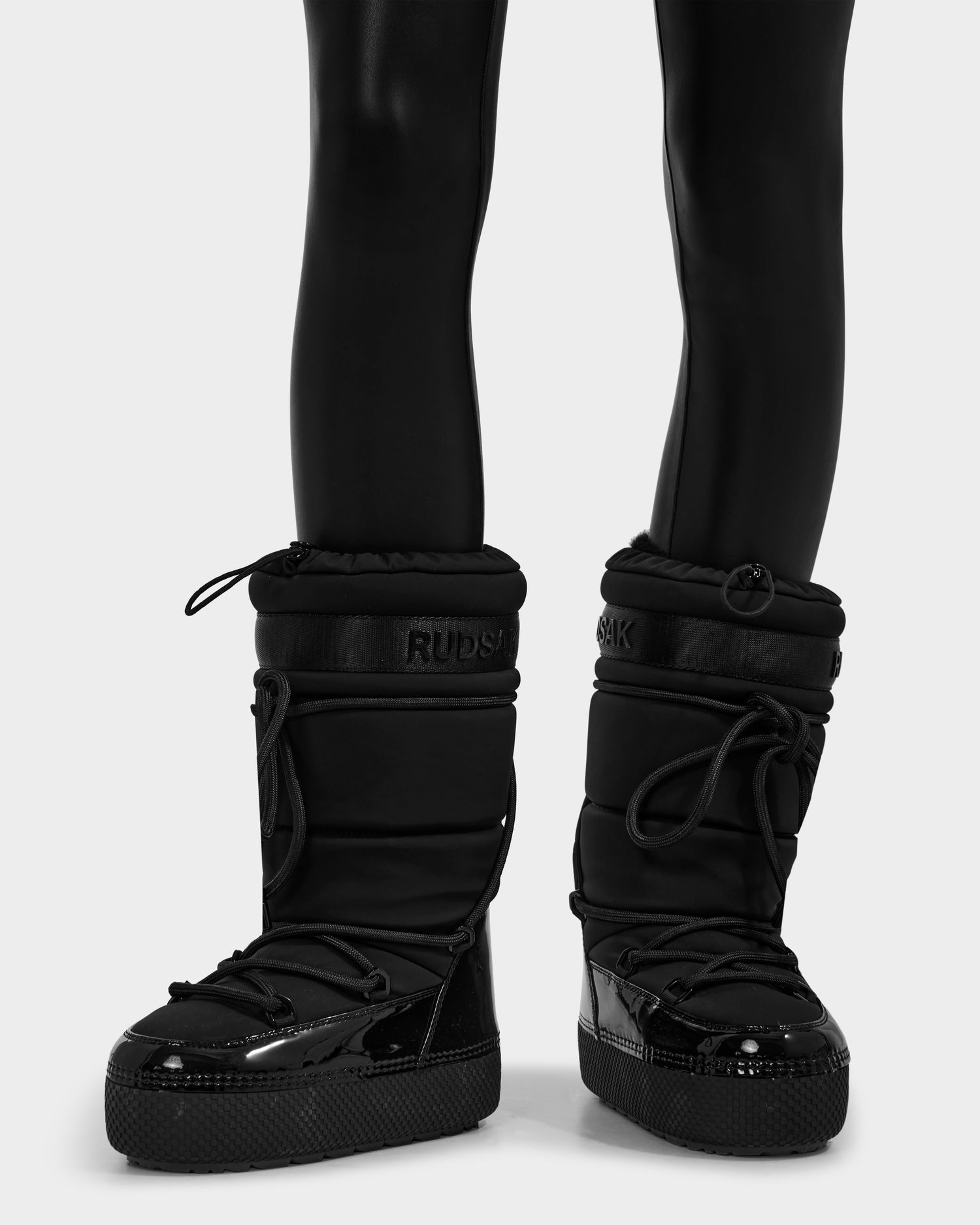 A person wearing Rudsak FARAH L - 8224542 Black shiny black leggings and tall moon boots with thick soles and drawstring closures, standing on a white background.