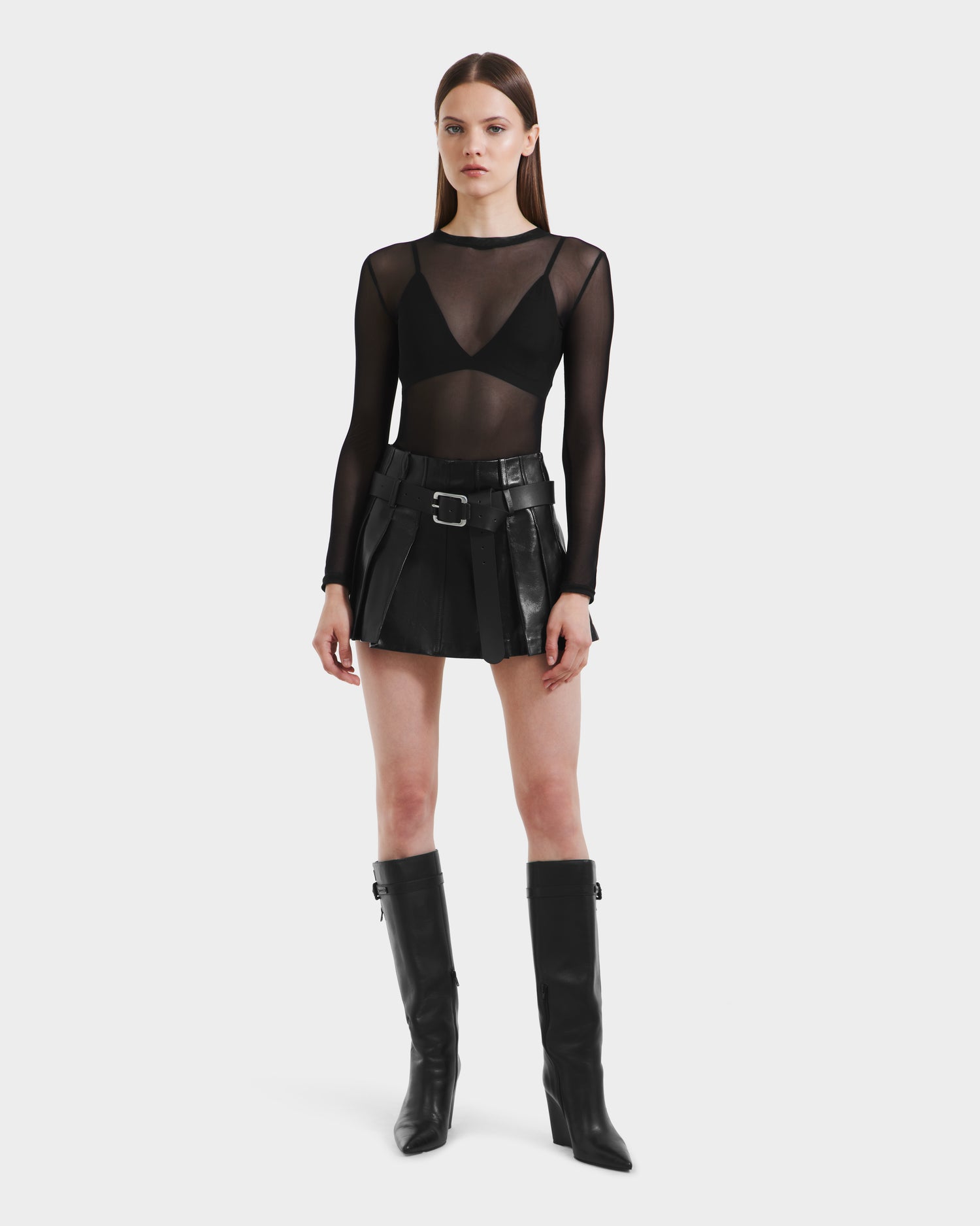 A woman poses against a plain background wearing the Rudsak FELICITY - 8525650 Black mini skirt with belt and buckle details, paired with a sheer black long-sleeve top, black bra, and knee-high black boots.
