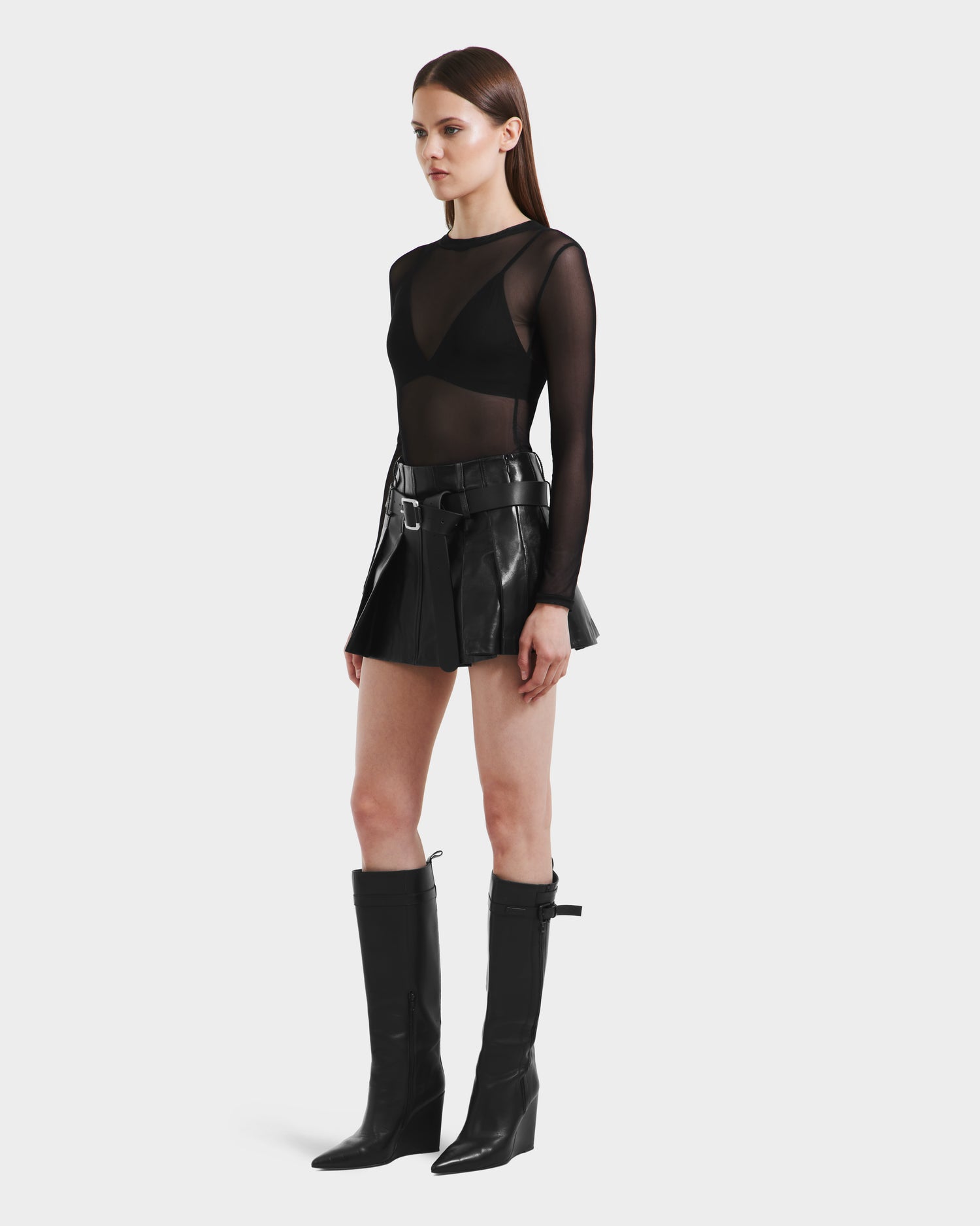A woman poses slightly to the side in Rudsaks FELICITY - 8525650 Black: a sheer black long-sleeve top, black bralette, and high-waist pleated leather skirt with belt, finished with knee-high black heeled boots against a white background.