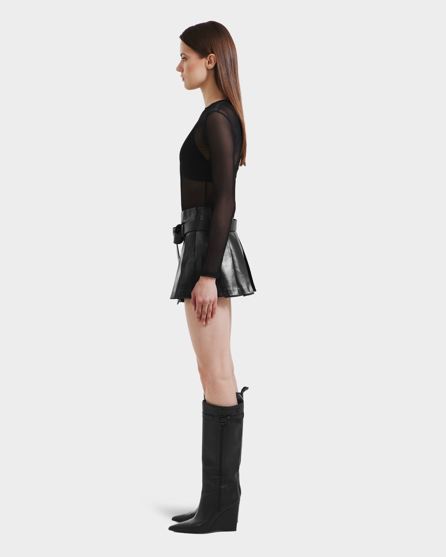 A woman stands in profile against a plain background, wearing the Rudsak FELICITY - 8525650 Black high-waist women’s leather mini skirt with a wide belt, paired with a sheer black long-sleeve top and knee-high black boots.