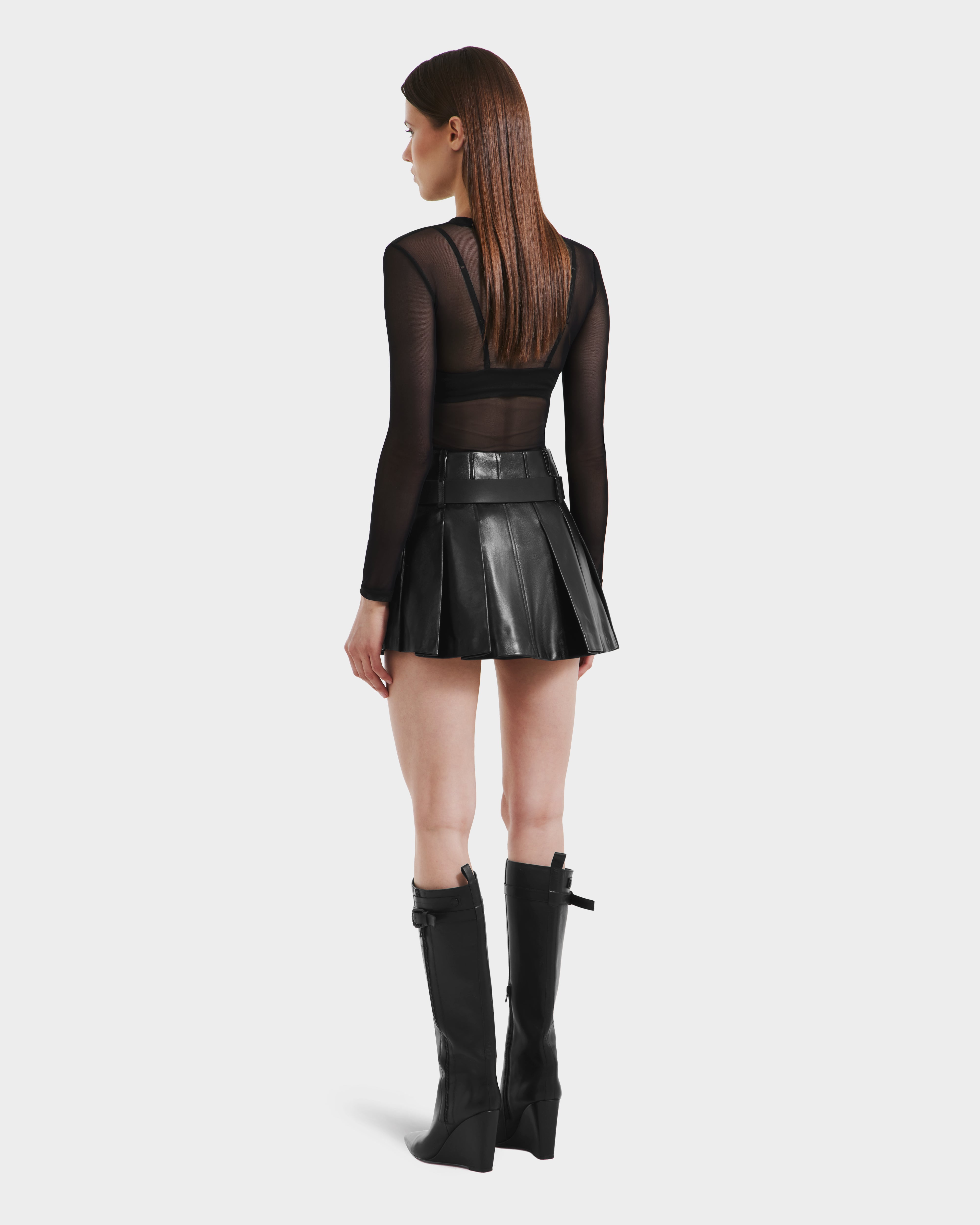 A woman with straight brown hair faces away, wearing Rudsaks FELICITY - 8525650 Black sheer long-sleeve top, a pleated leather skirt, and knee-high black boots against a plain light background.