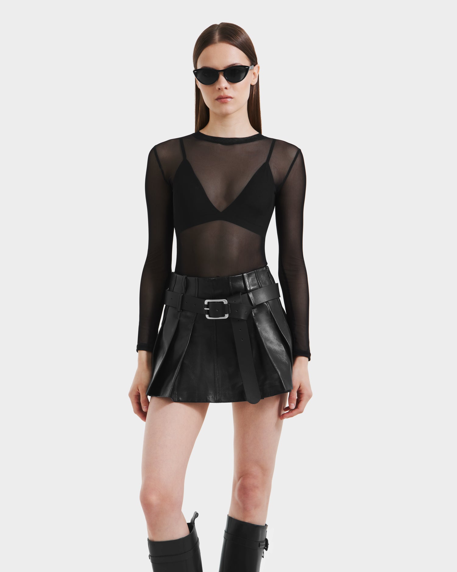 A woman models the Rudsak FELICITY - 8525650 Black leather mini skirt with a wide belt, paired with black sunglasses, a sheer long-sleeve top over a black bra, and knee-high boots against a plain white background.