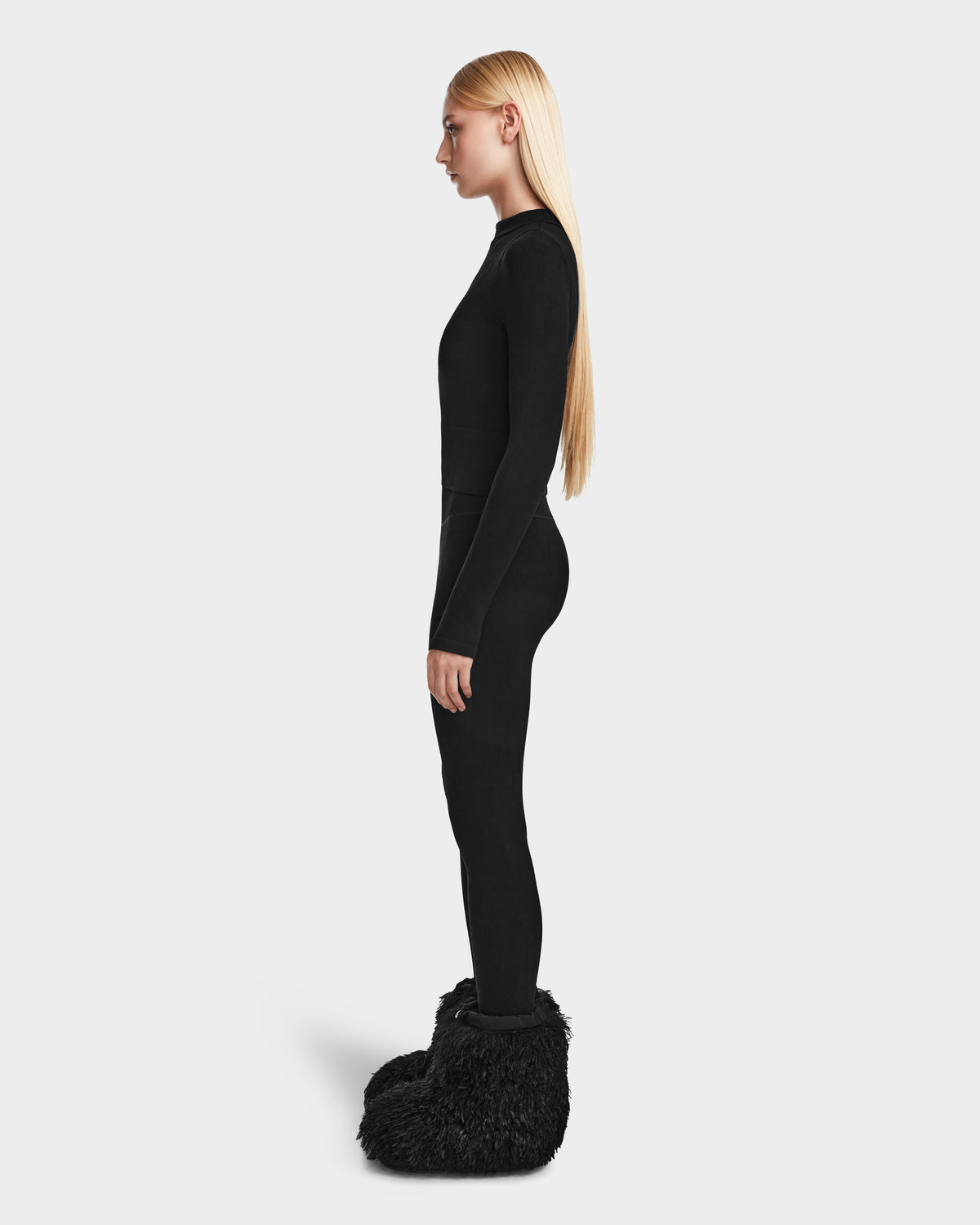 A woman with long blonde hair models the Rudsak FERKA - 8524542 Monogram Print Black boots with a fitted black long-sleeve top and pull-on leggings, creating an effortlessly chic winter look against a plain light background.