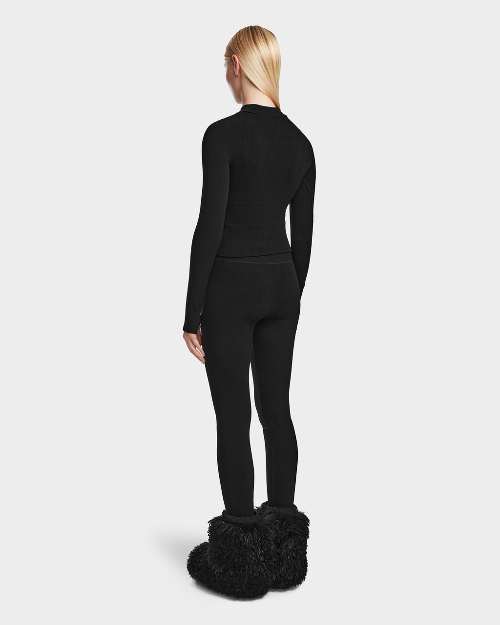 A person with straight blonde hair, seen from behind, wears the Rudsak FERKA - 8524542 Monogram Print Black fitted long-sleeve top and women’s pull-on leggings, paired with oversized black furry slippers against a plain light background.