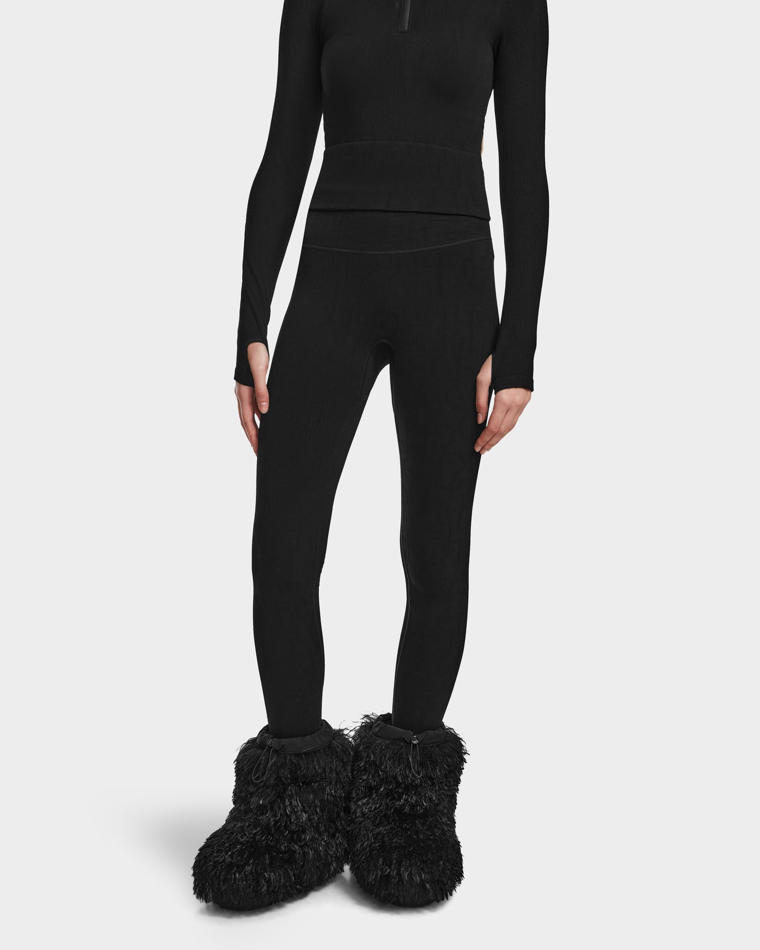 A person models the Rudsak FERKA - 8524542 Monogram Print Black in an all-black winter look with a long-sleeve top, thermal leggings, and oversized fluffy black slippers against a plain white background; their head is not visible.