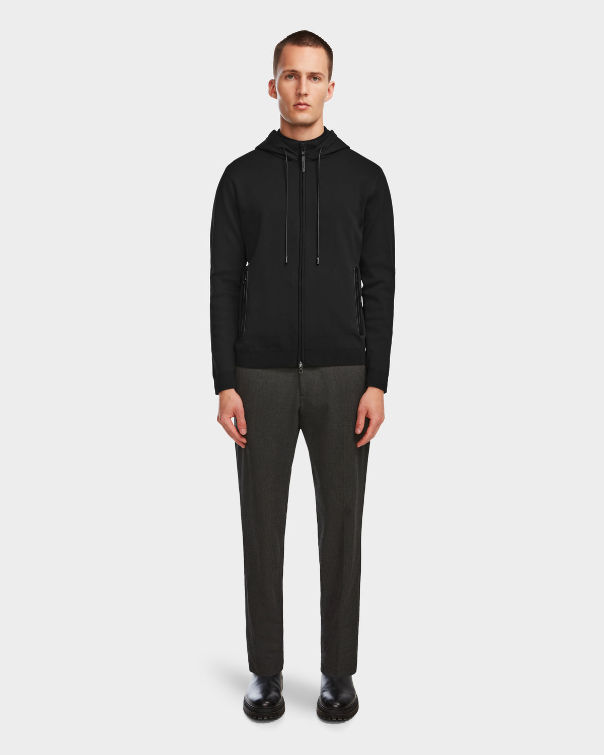 A man faces forward against a plain white background, wearing the Rudsak FILEY - 6524512 Black mens knit hoodie, along with dark gray trousers and black shoes.