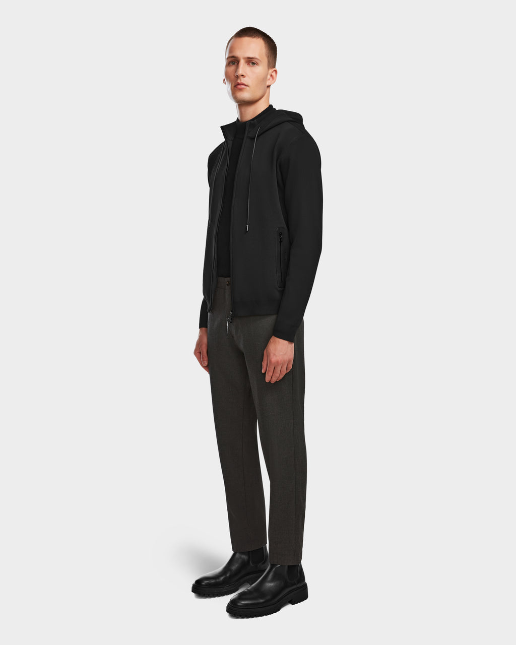 A man stands slightly turned, wearing the Rudsak FILEY - 6524512 Black knit hoodie with dark gray pants and black boots, set against a plain light gray background.