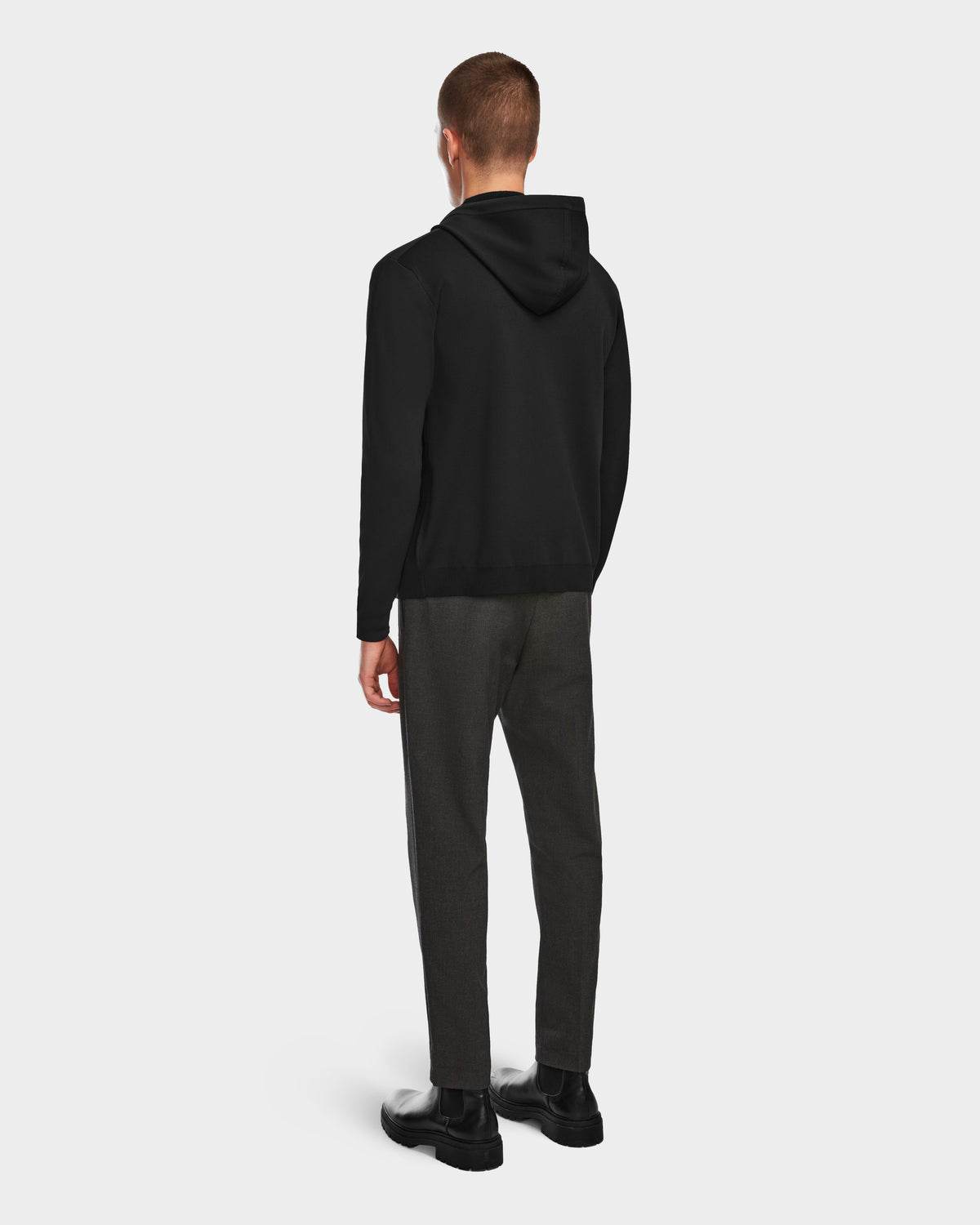 A person faces away from the camera, wearing Rudsaks FILEY - 6524512 Black hoodie, gray pants, and black shoes against a plain light gray background.