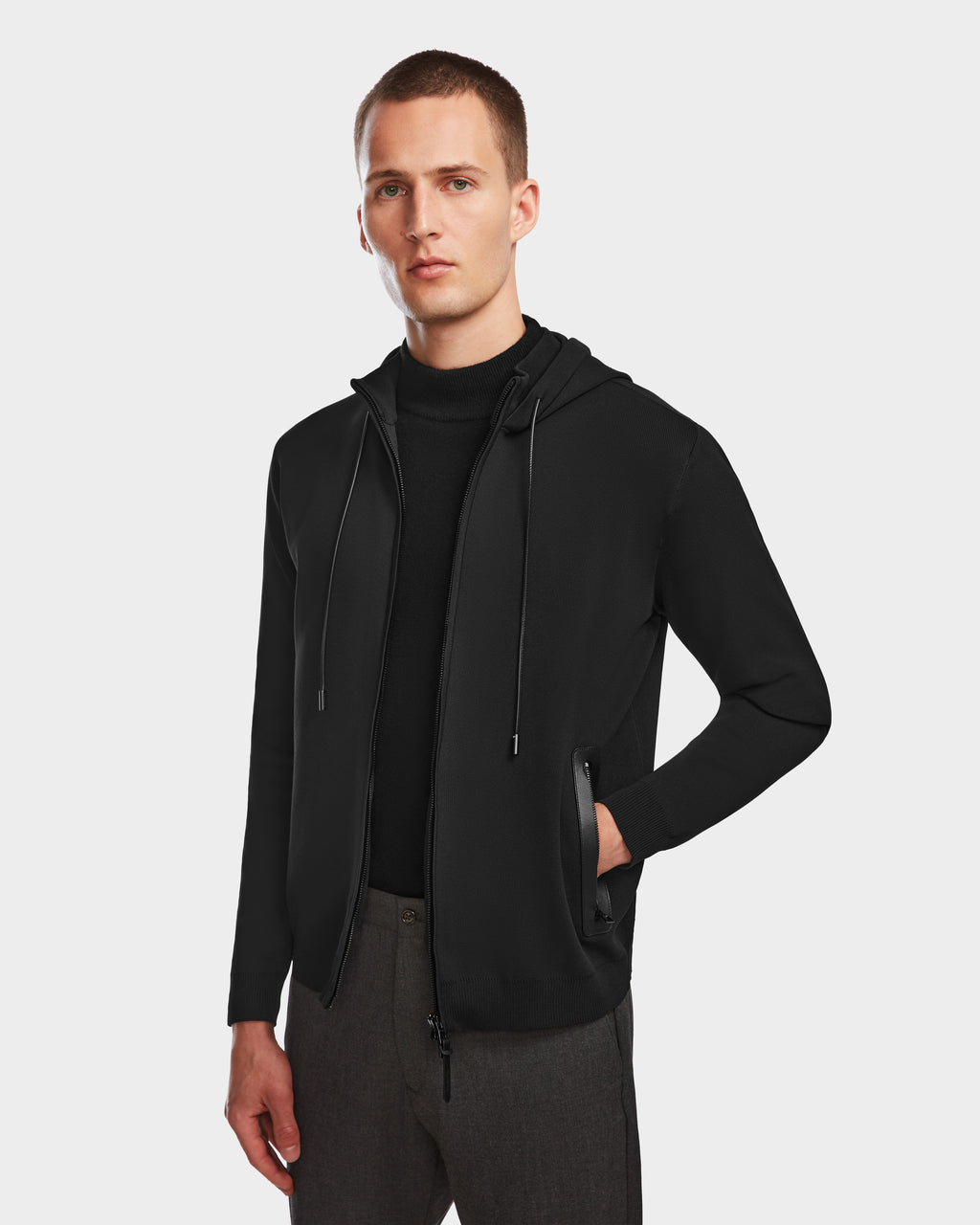 A man with short hair wears the Rudsak FILEY - 6524512 Black full-zip hoodie over a black shirt and dark trousers, standing against a plain light background with one hand in his pocket.