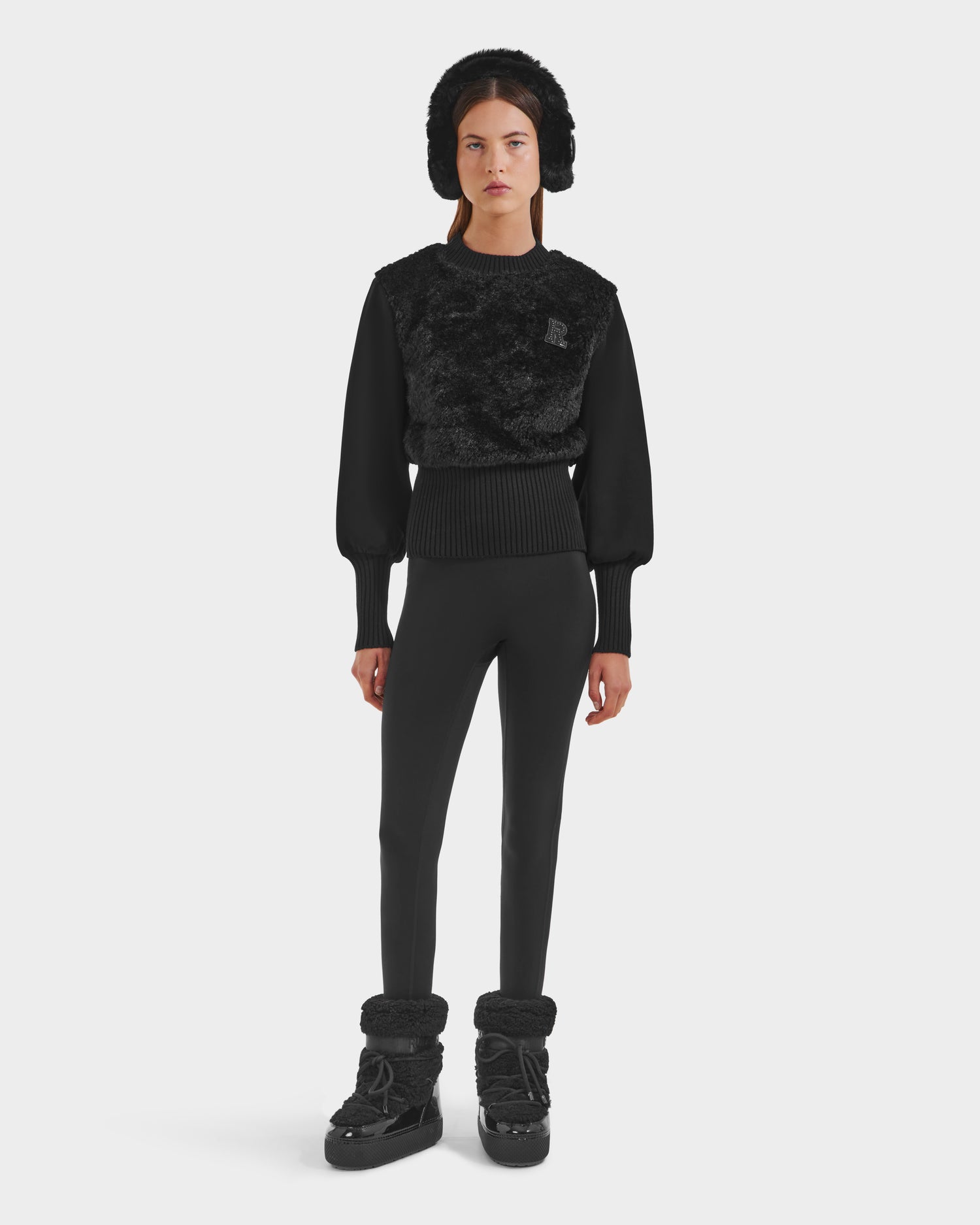 A woman models the FLORENCE - 8525546 Black by Rudsak: all-black winter wear, including a textured sweater, fitted pants, après-ski top, winter boots, and a fur-lined hat. She stands against a white background with cozy elegance.