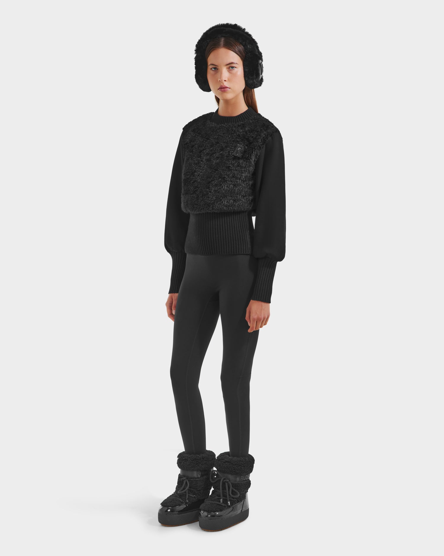A woman faces forward against a white background, wearing Rudsak’s FLORENCE - 8525546 Black boots, paired with a textured sweater and fitted leggings for a chic, all-black winter look.
