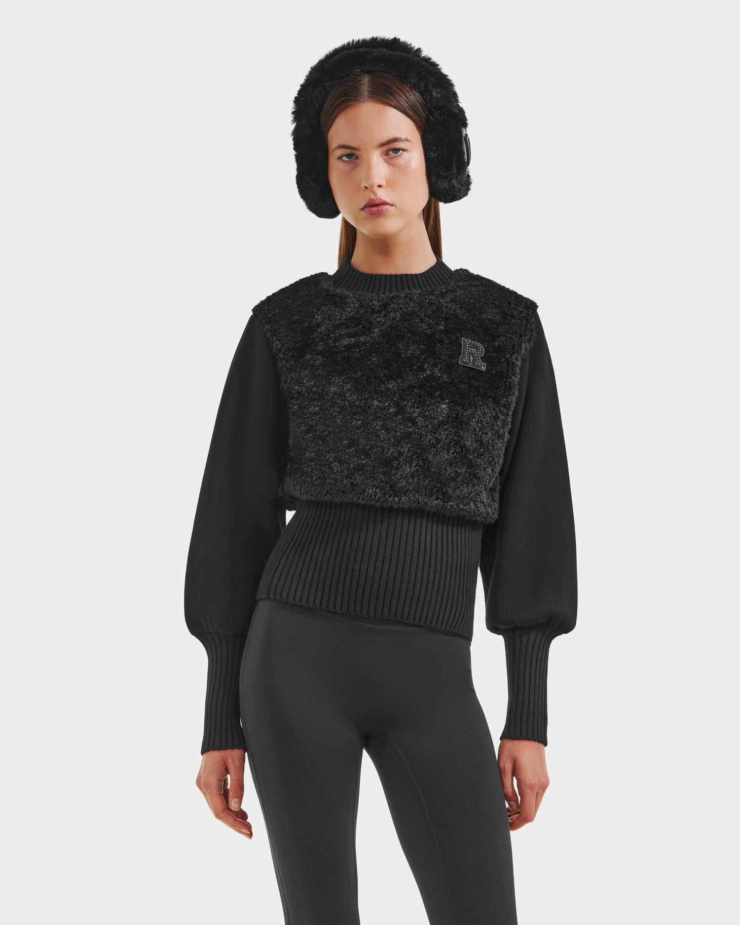 A woman stands against a plain light background wearing the Rudsak FLORENCE - 8525546 Black faux fur women's top and leggings, her cozy glamorous look finished with a textured sweater over a ribbed puff-sleeve top.