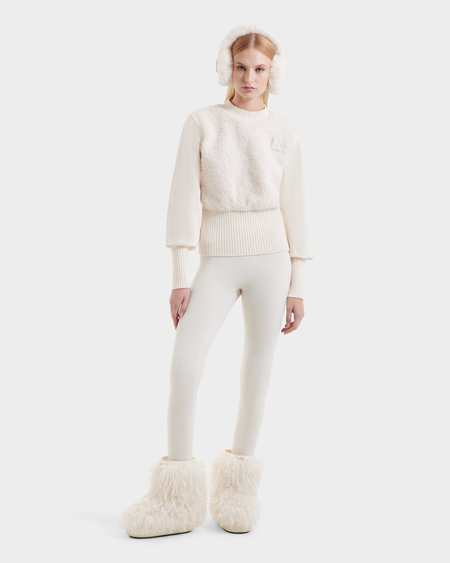 A woman wearing the Rudsak FLORENCE - 8525546 Iceland stands in all-white winter attire—textured sweater, earmuffs, fitted leggings, and fluffy boots—against a plain white background.