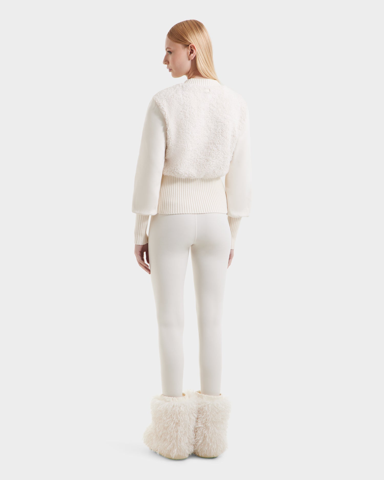 A woman with straight blonde hair stands facing away, embracing cozy glamour in the Rudsak FLORENCE - 8525546 Iceland textured off-white sweater, fitted white pants, and fluffy white boots—perfect après-ski essentials.