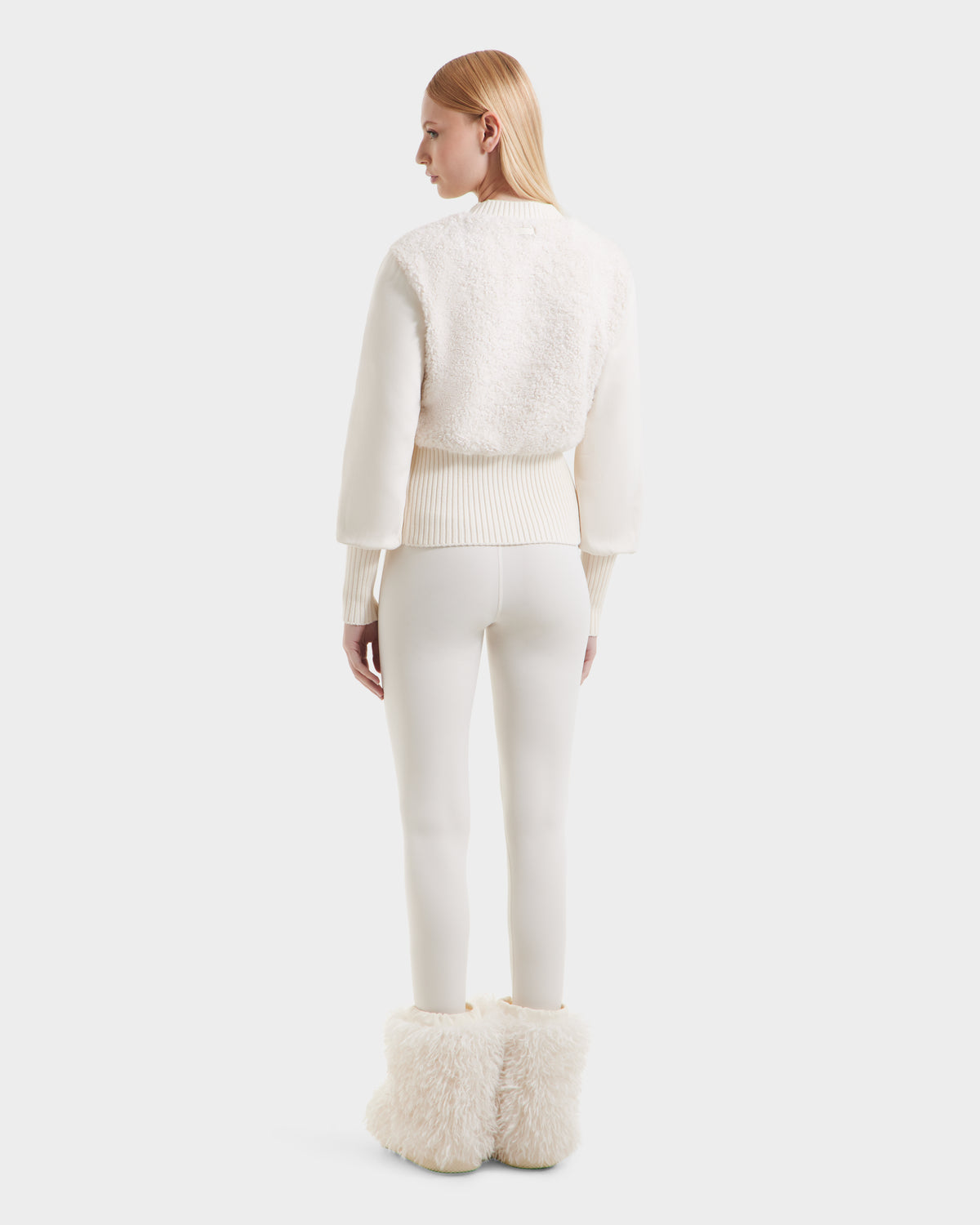 A woman with straight blonde hair stands facing away, embracing cozy glamour in the Rudsak FLORENCE - 8525546 Iceland textured off-white sweater, fitted white pants, and fluffy white boots—perfect après-ski essentials.
