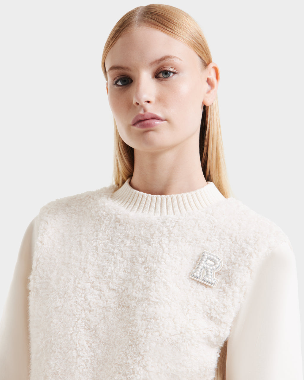 A woman with straight blonde hair channels cozy glamour in the Rudsak FLORENCE - 8525546 Iceland off-white textured sweater with a ribbed collar and embellished letter R brooch, capturing modern après-ski style against a plain background.