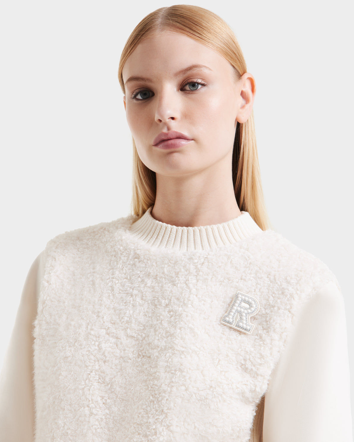 A woman with straight blonde hair channels cozy glamour in the Rudsak FLORENCE - 8525546 Iceland off-white textured sweater with a ribbed collar and embellished letter R brooch, capturing modern après-ski style against a plain background.