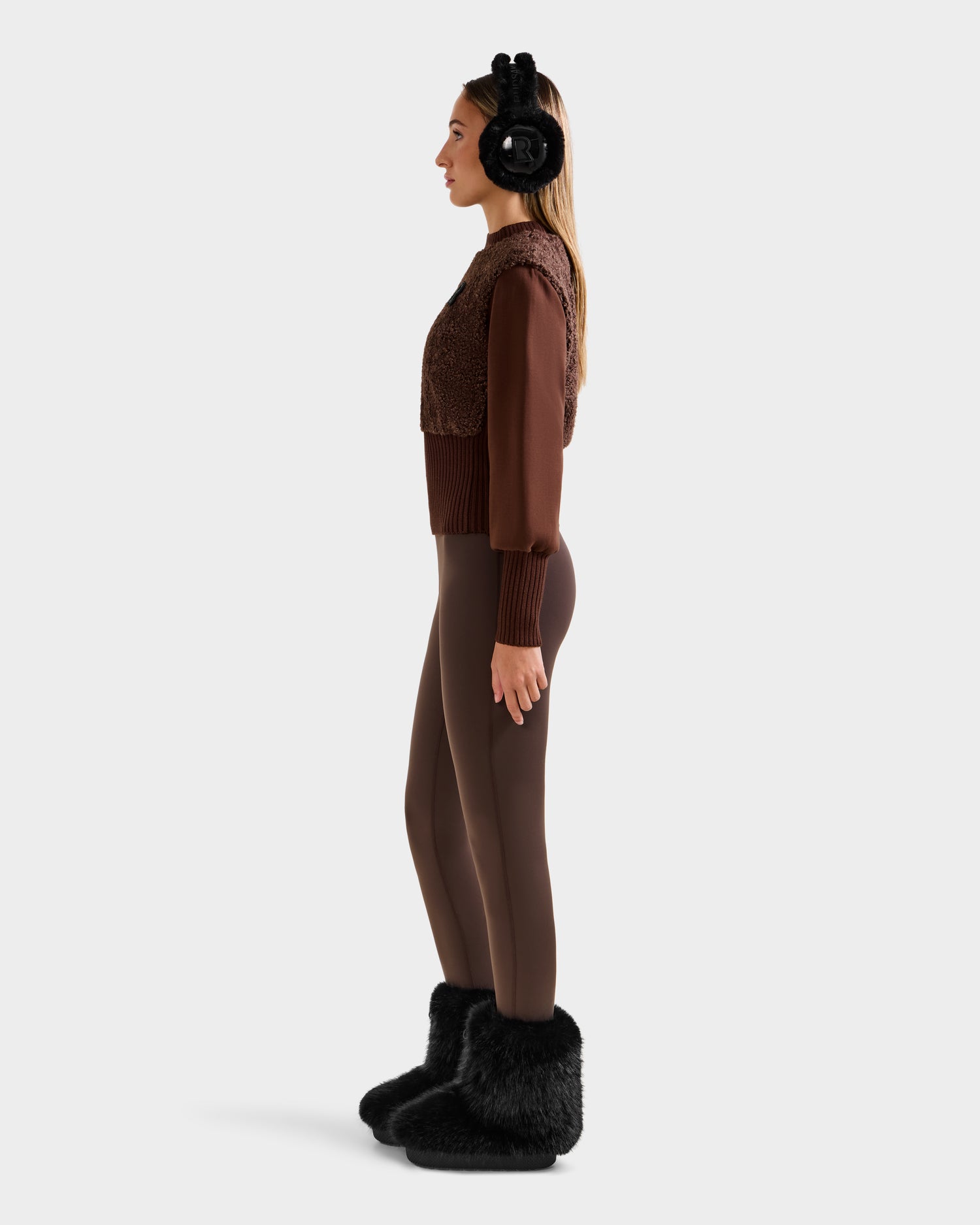 A woman in profile wears Rudsaks FLORENCE - 8525546 Mocha top, styled with brown leggings, black earmuffs, and fuzzy slippers, radiating cozy glamour against a plain light background.