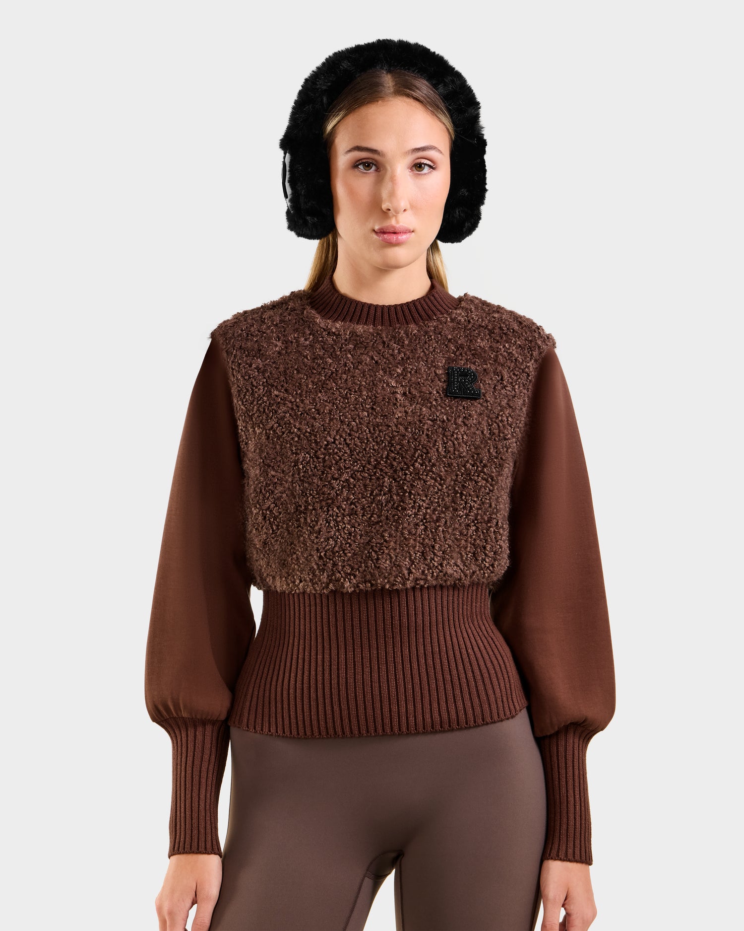 A woman channels après-ski glamour in Rudsak’s FLORENCE - 8525546 Mocha leggings and a textured sweater, accessorized with black earmuffs, set against a simple light background.