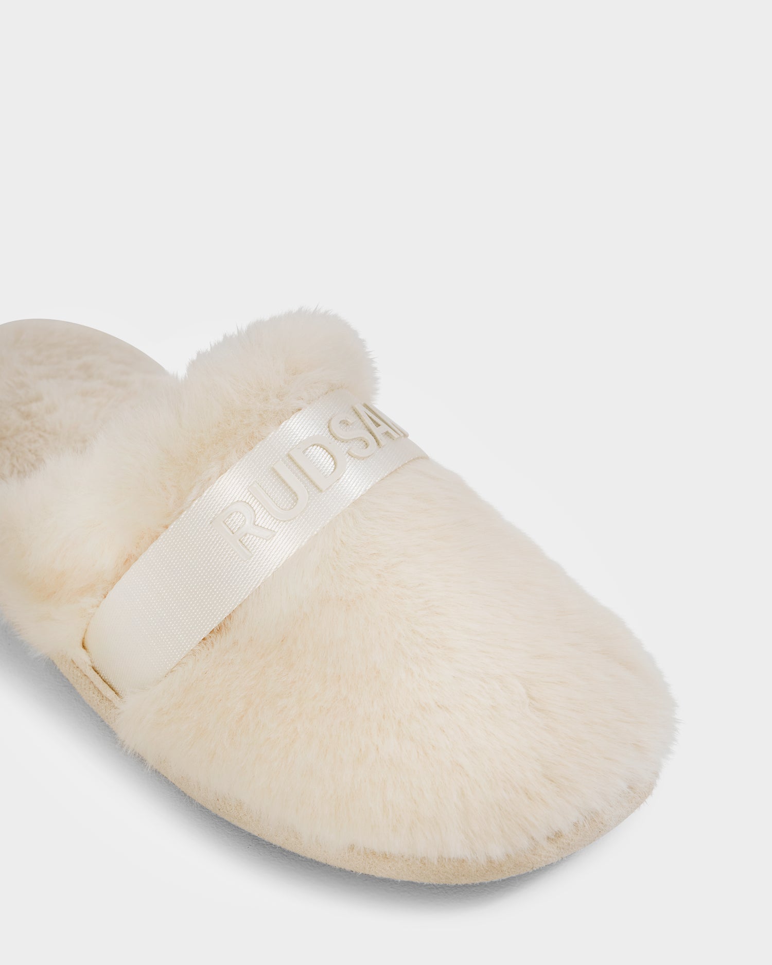 A close-up of the FLUFF - L223509 Cream women's slipper by Rudsak, featuring cream-colored faux fur and a wide shiny band with embossed RUDSAK text, set against a plain white background for a cozy lounge look.