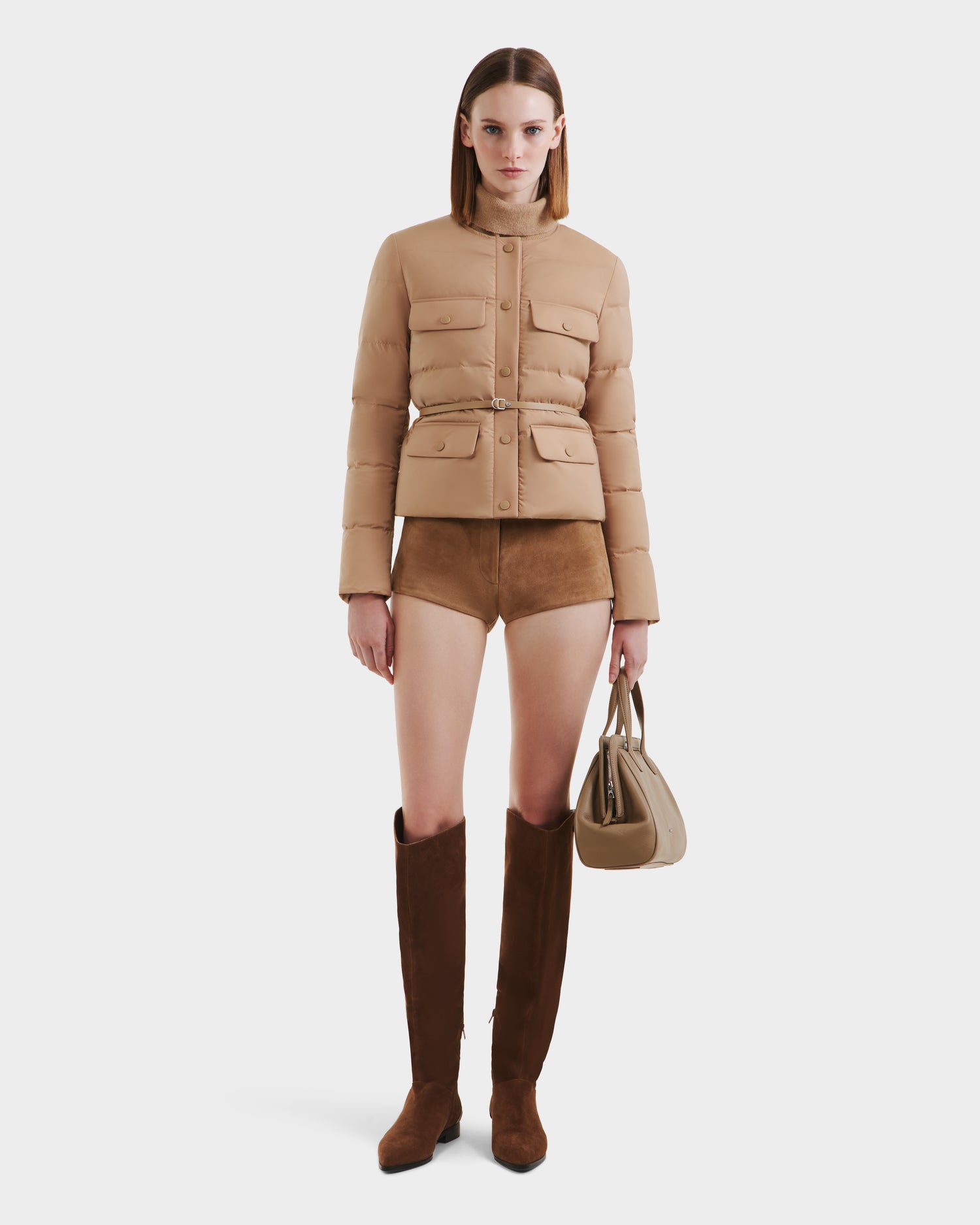 A woman stands against a white background wearing the Rudsak FRANCESCA - 8125021 Almond RDS-certified down belted puffer jacket, brown suede shorts, tall brown suede boots, and holding a beige handbag.