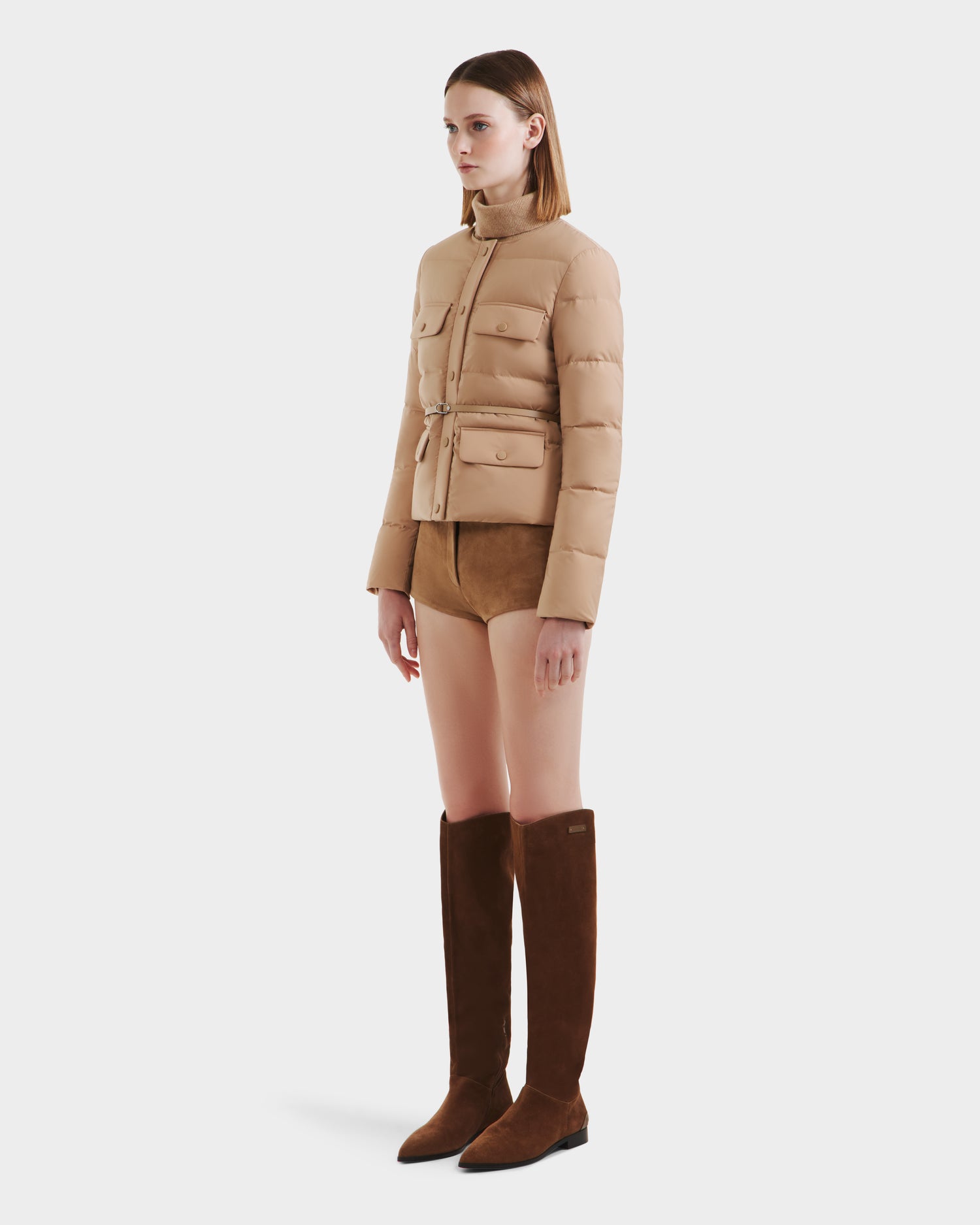 A woman faces left against a white background, wearing the Rudsak FRANCESCA - 8125021 Almond beige quilted puffer jacket with buttoned pockets, a vegan leather belt, tan suede shorts, and tall brown suede boots.