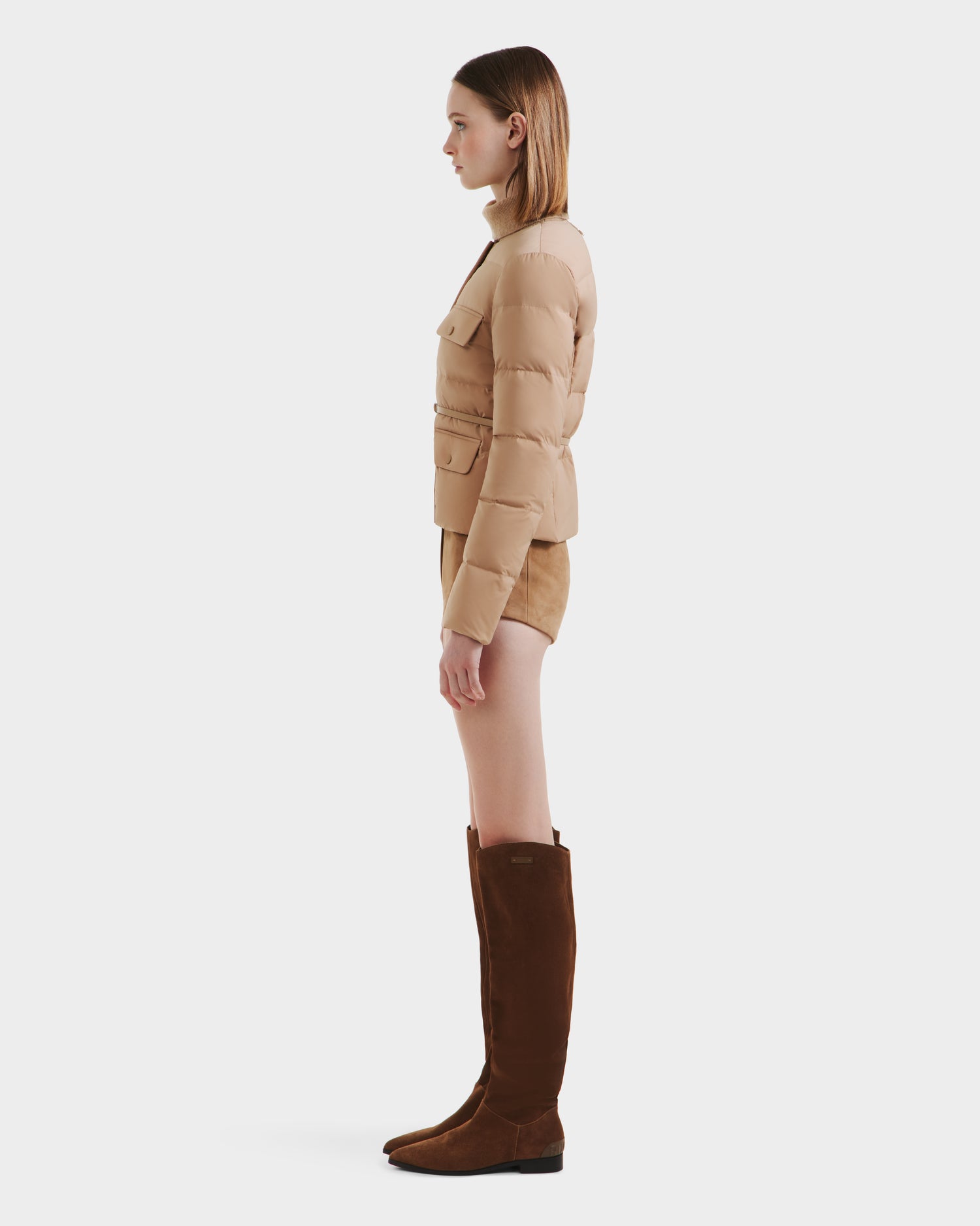 A woman stands in profile wearing the Rudsak FRANCESCA - 8125021 Almond quilted puffer jacket, tan shorts, and tall brown suede boots against a plain white background.