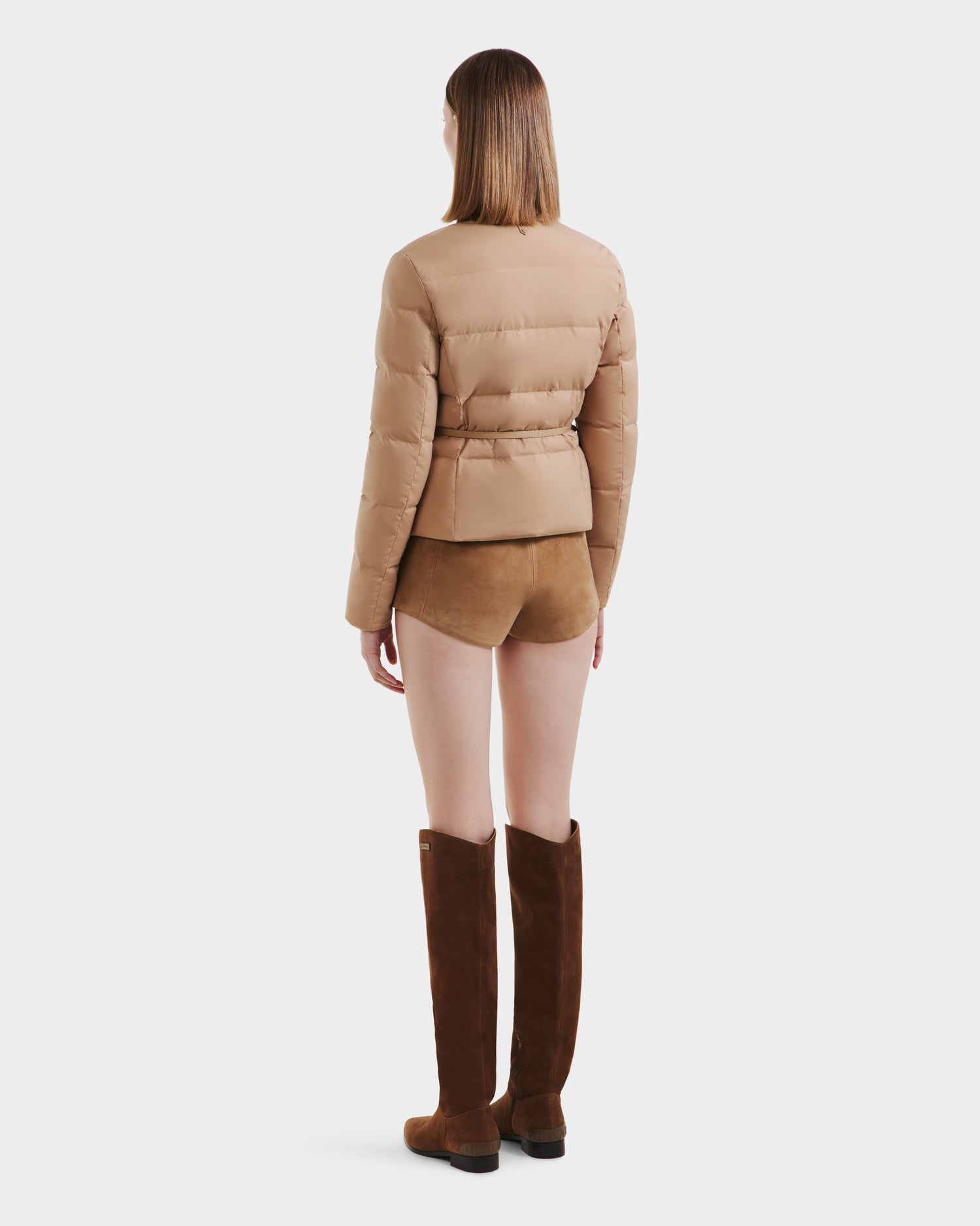 A woman, posed against a plain white background, faces away wearing the Rudsak FRANCESCA - 8125021 Almond quilted puffer jacket. She pairs it with tan suede shorts, a vegan leather belt, knee-high brown suede boots, and has straight shoulder-length hair.