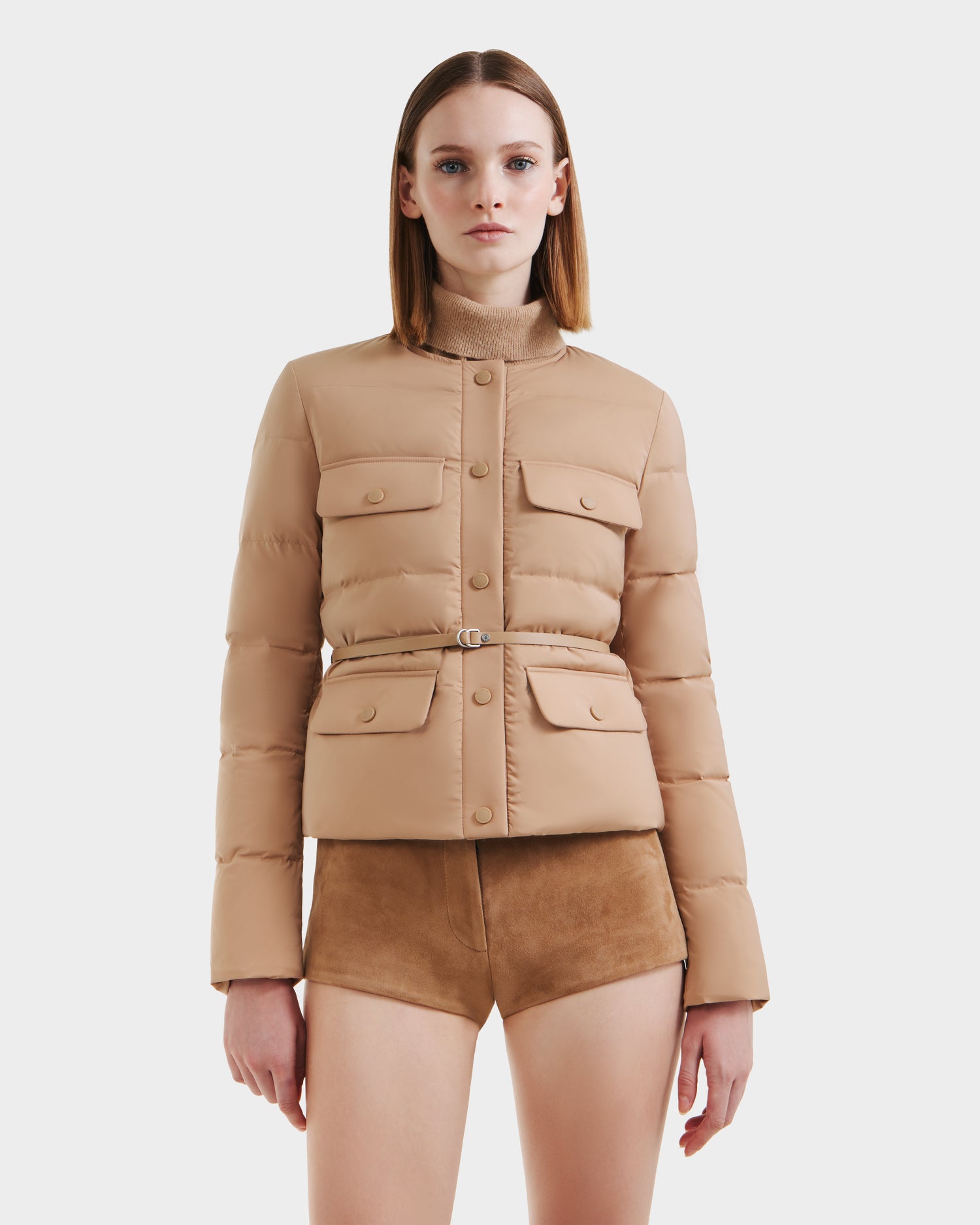 A woman with straight, shoulder-length hair models the Rudsak FRANCESCA - 8125021 Almond quilted puffer jacket with front pockets and brown suede shorts, standing against a plain white background.