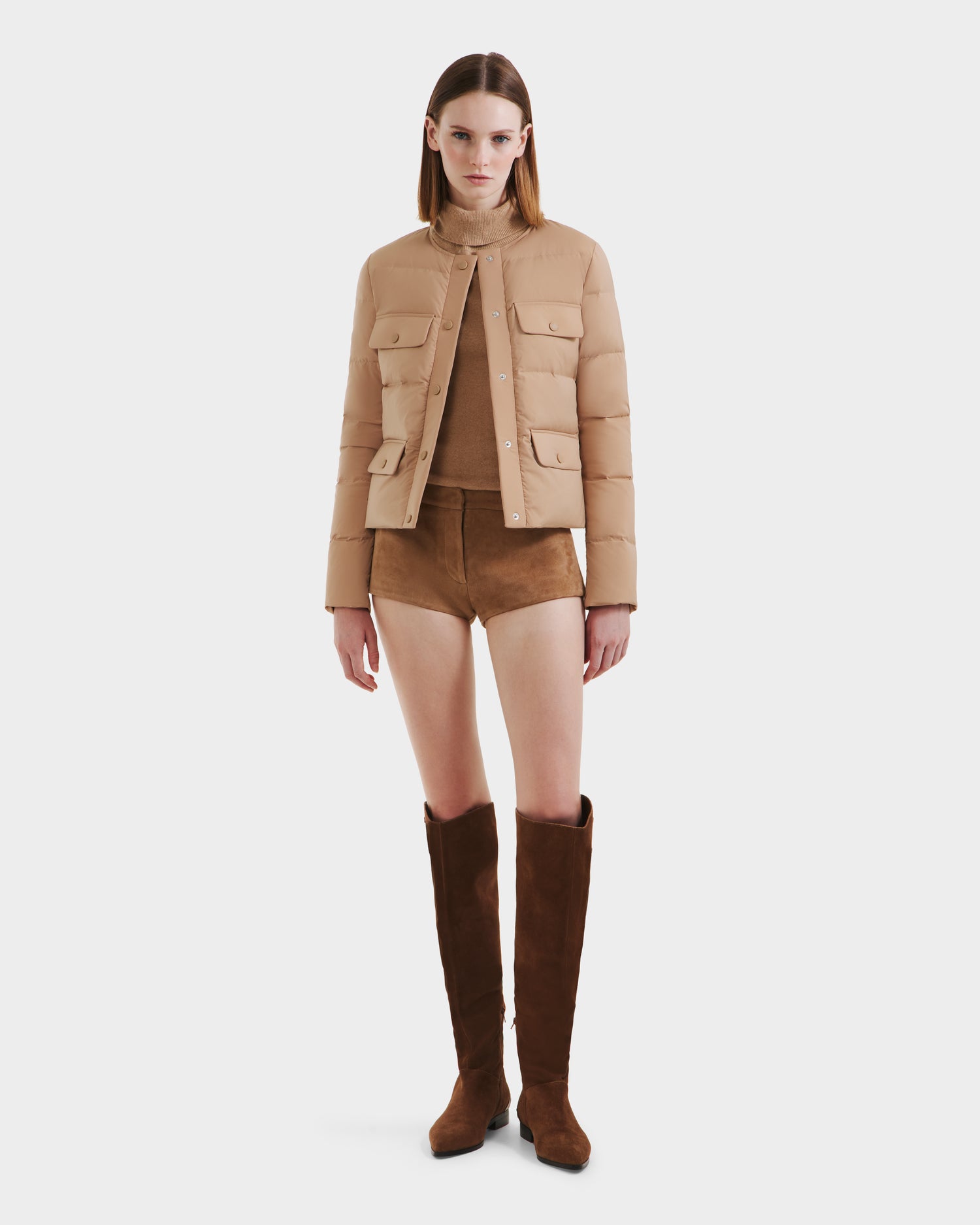 A woman stands facing forward against a white background, wearing the Rudsak FRANCESCA - 8125021 Almond beige quilted puffer jacket with RDS-certified down, a tan top, brown suede shorts, and knee-high brown suede boots.