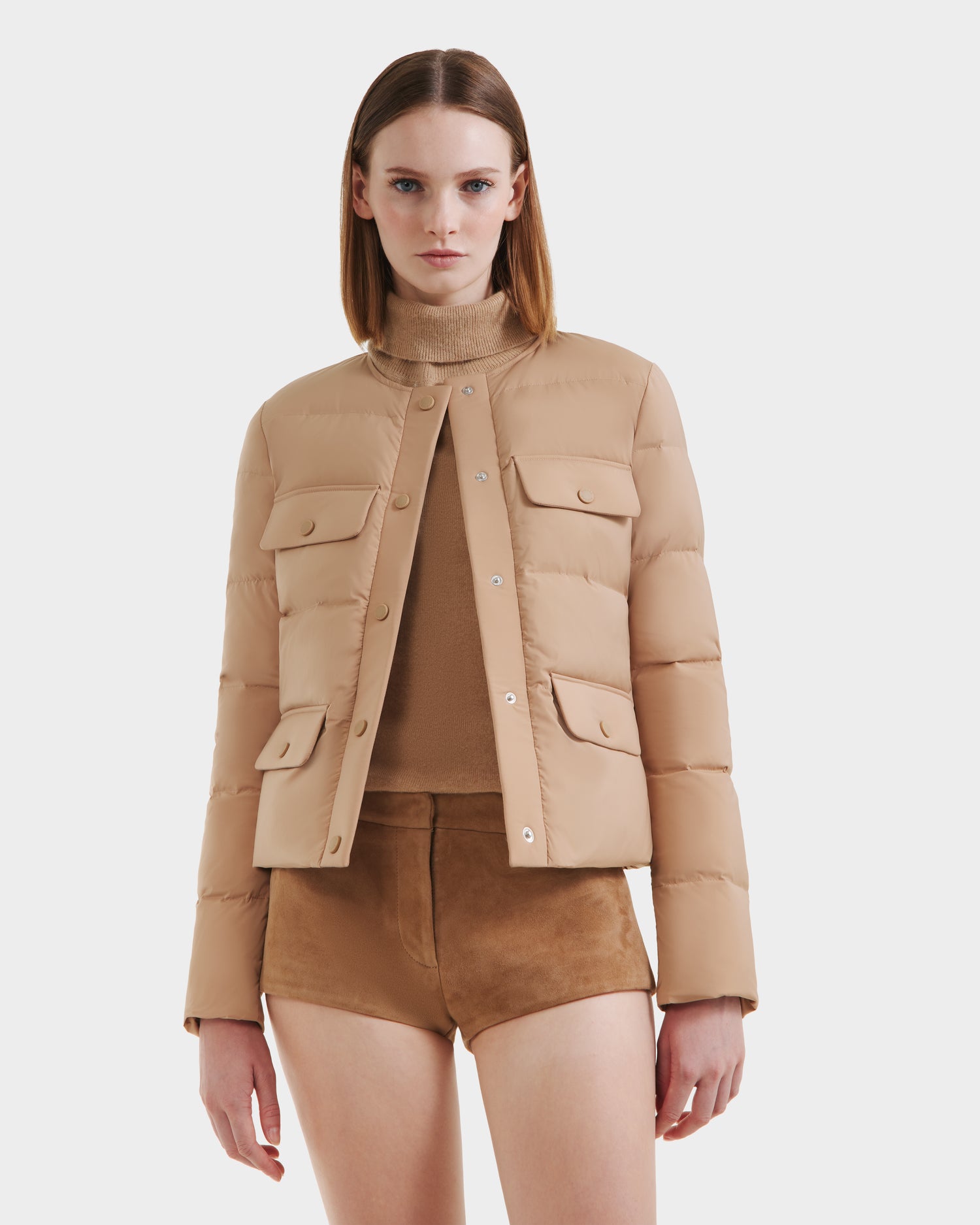 A woman wears the Rudsak FRANCESCA - 8125021 Almond puffer jacket over a beige turtleneck and brown suede shorts with a vegan leather belt, standing against a plain white background.