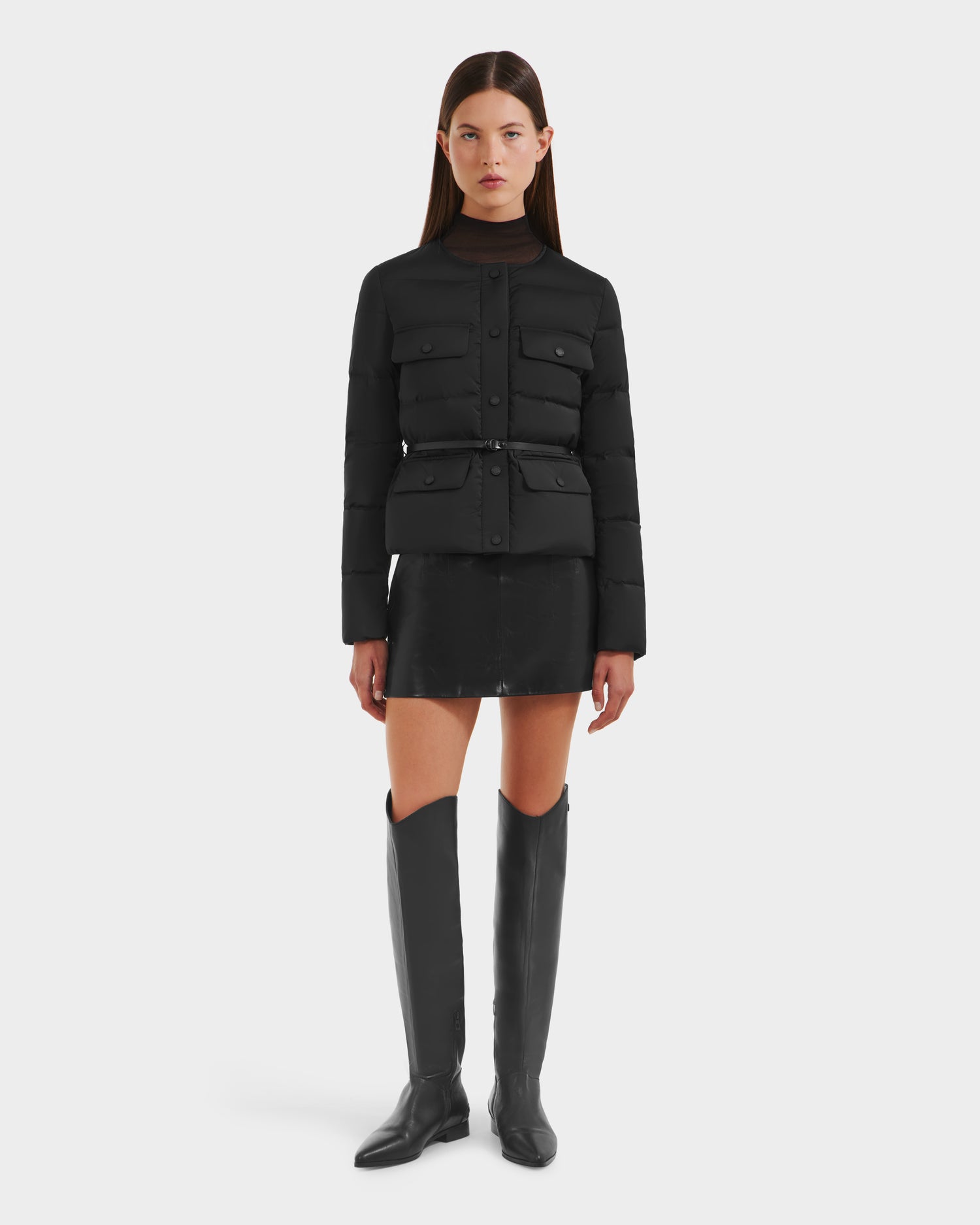 A woman with straight brown hair and a neutral expression faces forward against a white background, wearing Rudsak’s FRANCESCA - 8125021 Black lightweight puffer jacket with vegan leather belt, a black mini skirt, and knee-high boots.