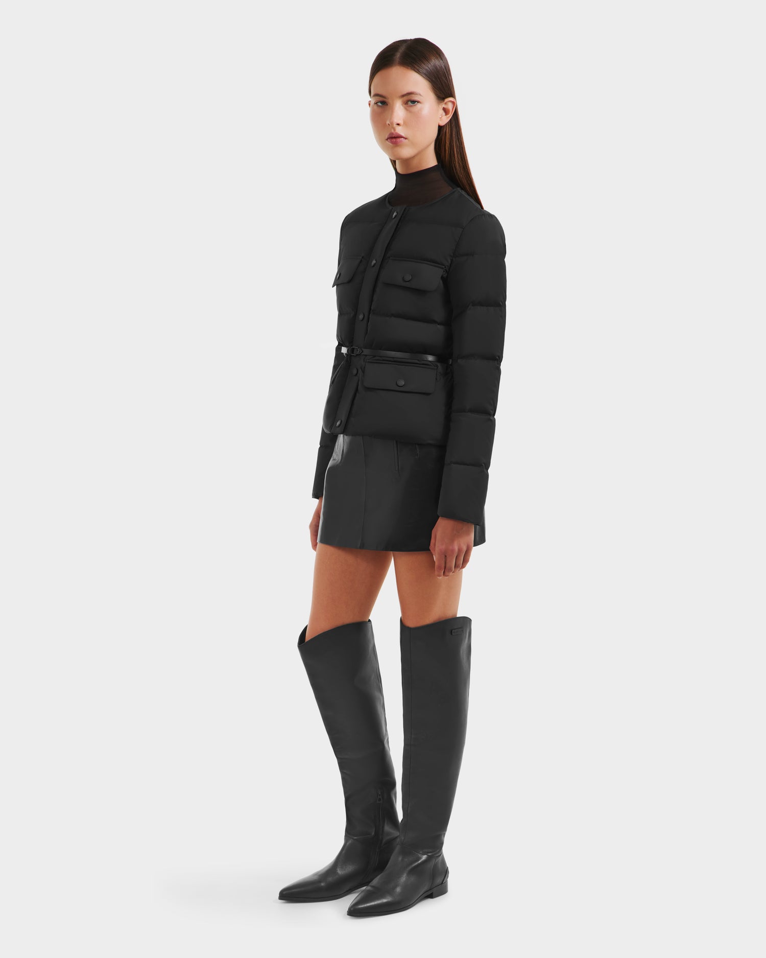 A woman stands against a plain background wearing the Rudsak FRANCESCA - 8125021 Black lightweight puffer jacket with front pockets, styled with a black mini skirt and tall black boots. Her hair is straight and she has a neutral expression.